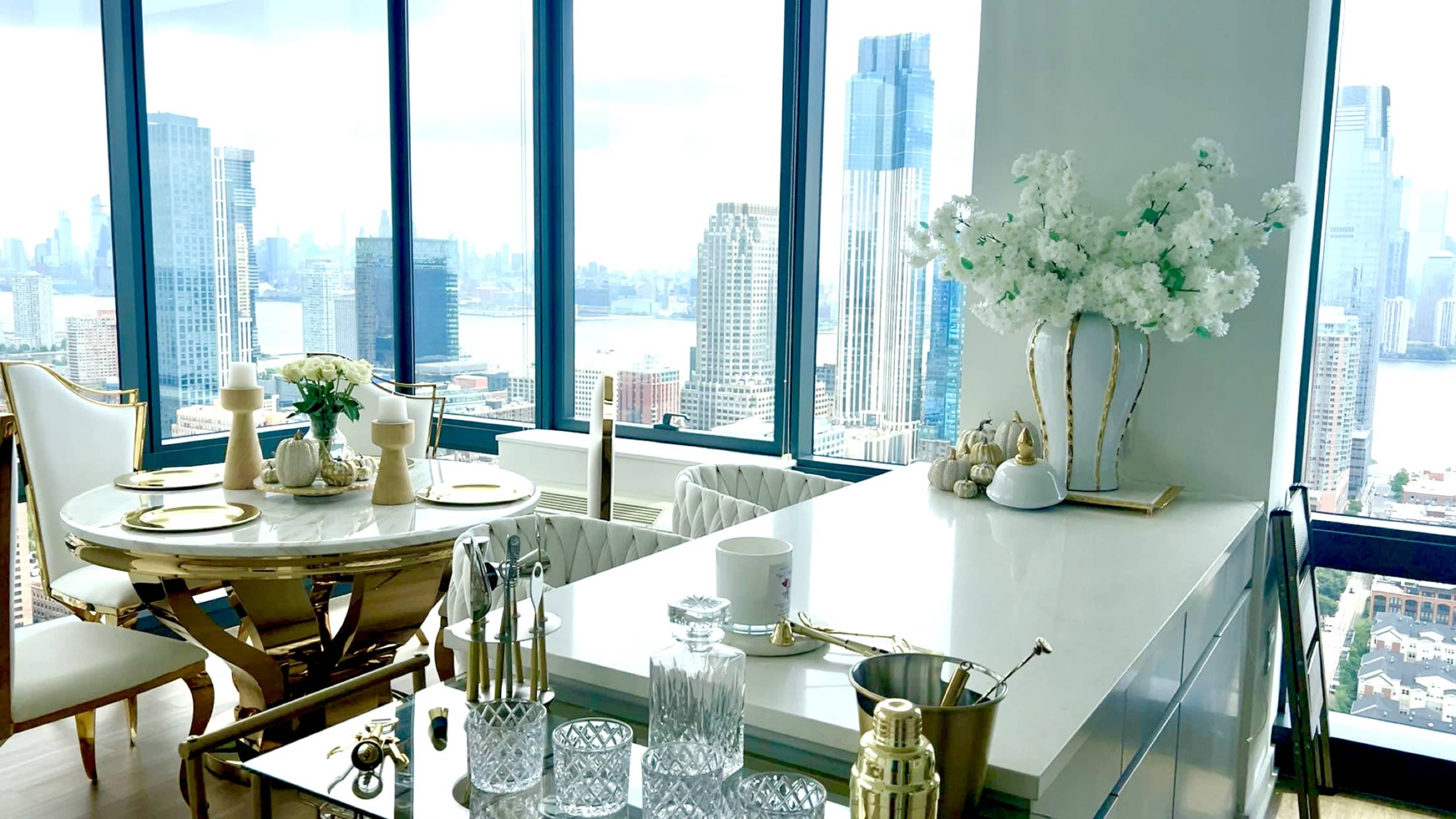 The image shows a modern dining area with a round table set for dining, surrounded by windows offering a view of a city skyline.