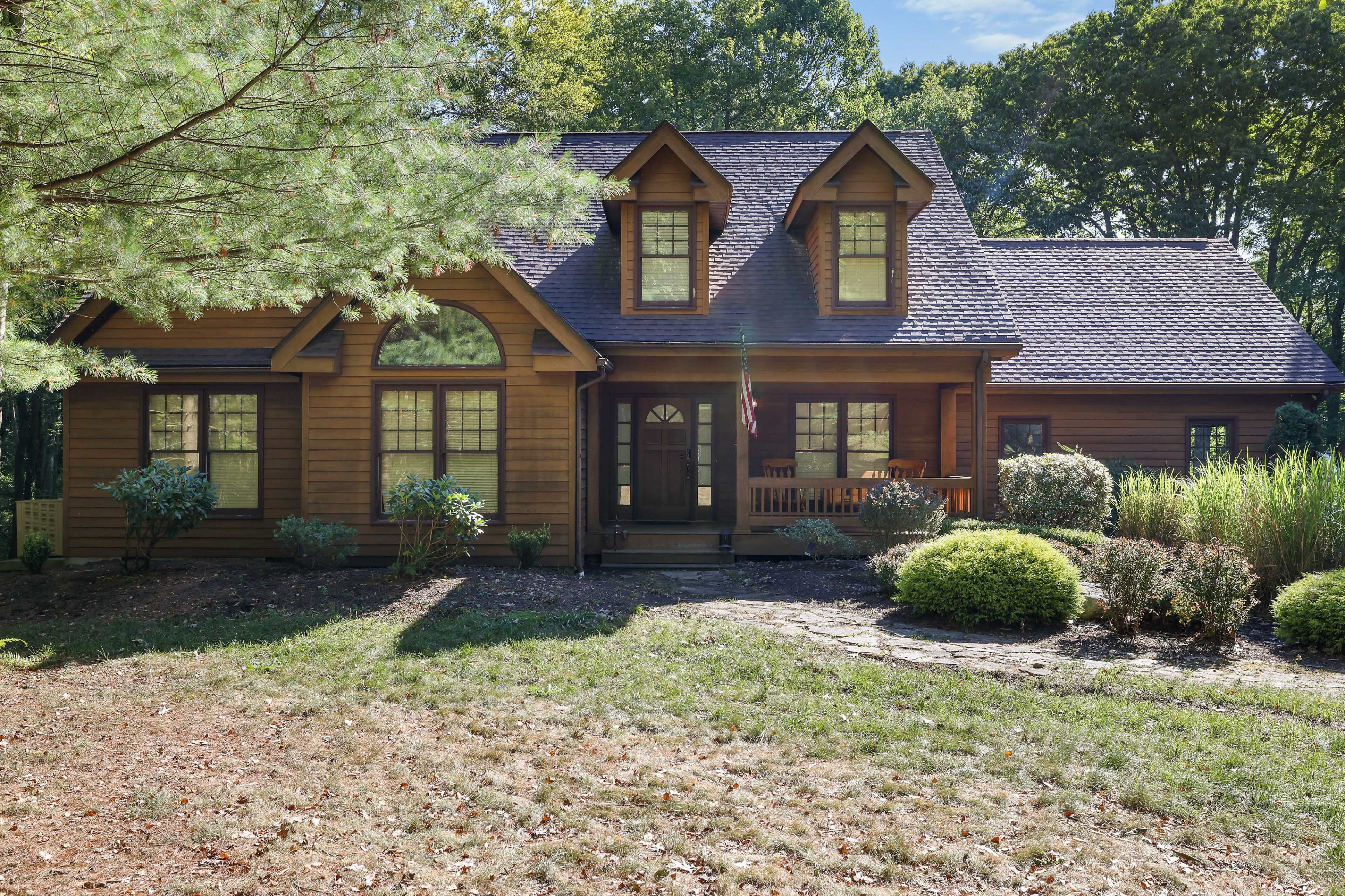 The image shows a large wooden house surrounded by green trees and shrubs, with a pathway leading to the front door.