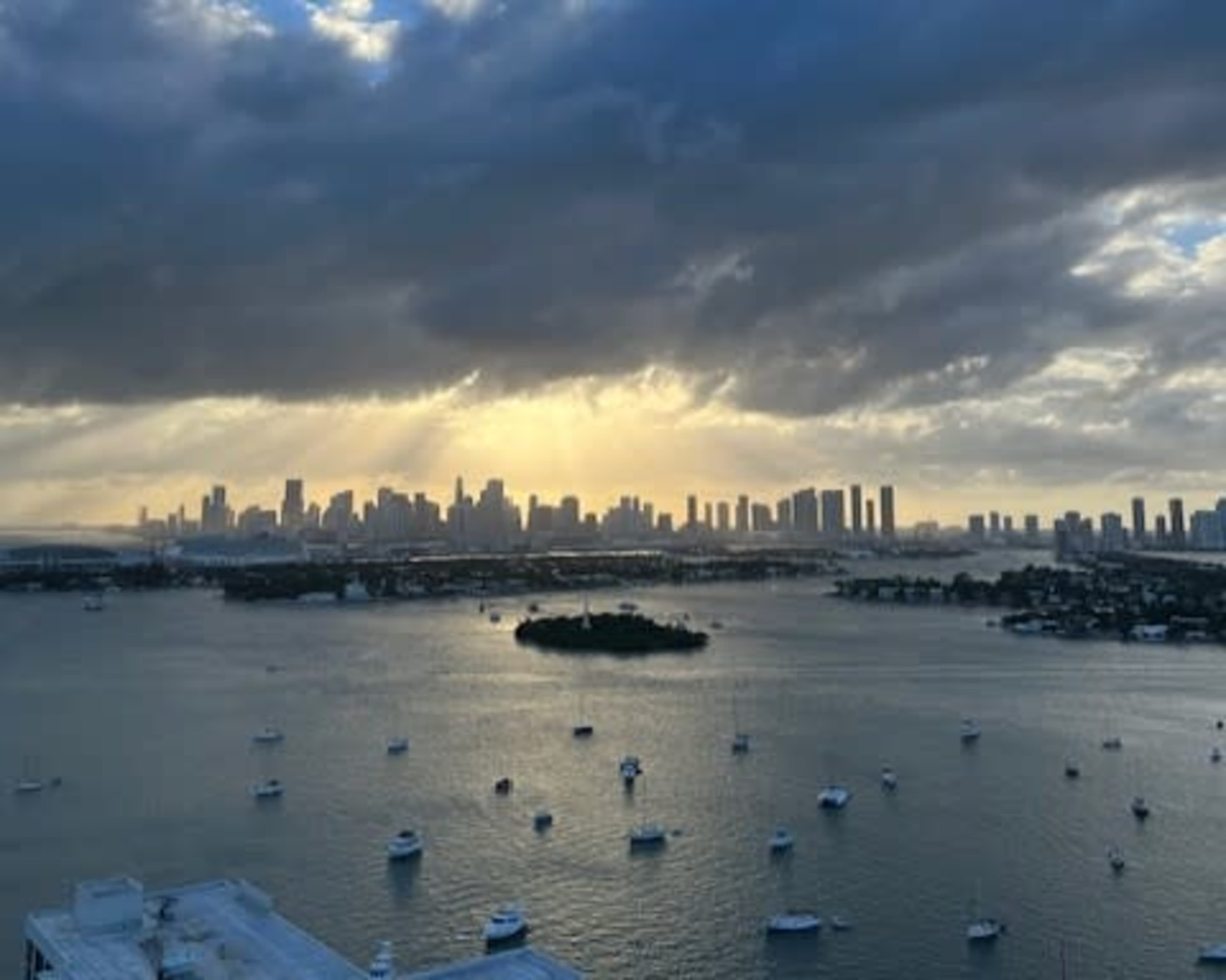 A city skyline is visible under a cloudy sky, with sunlight illuminating the buildings and reflecting off the water where boats are anchored.