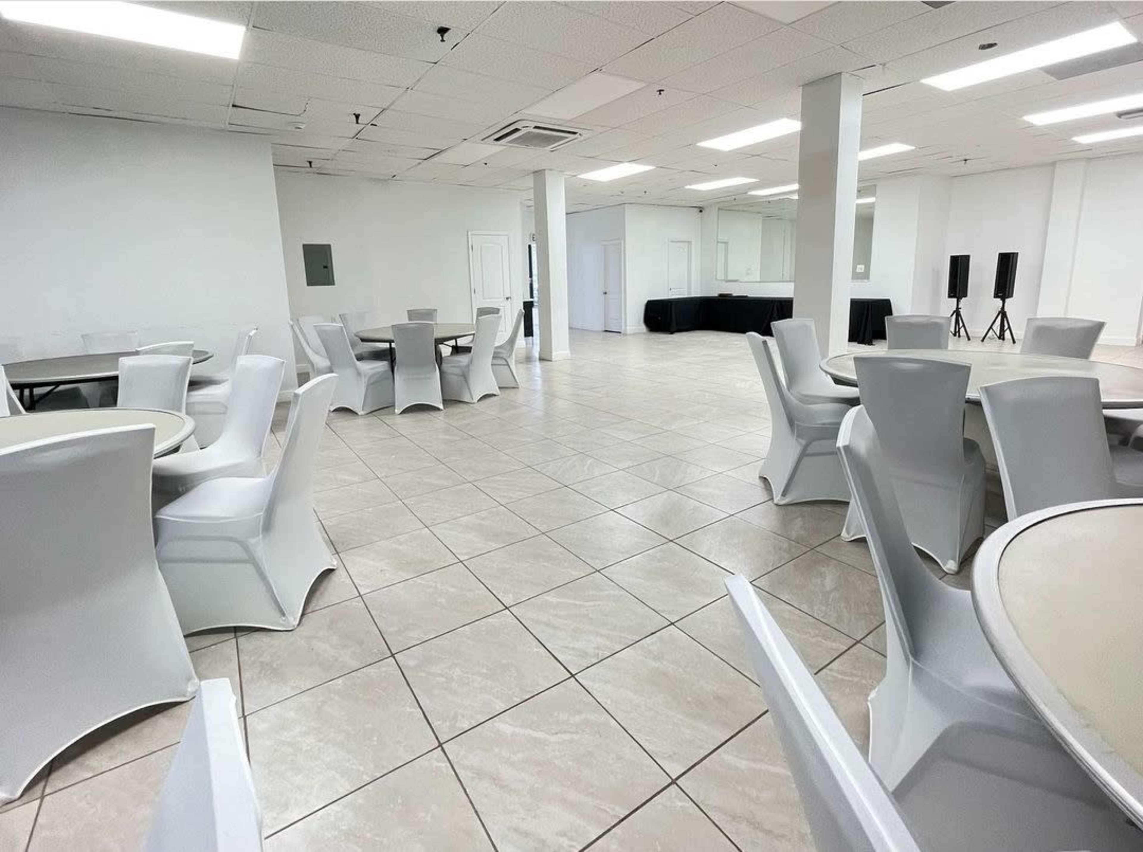 The image shows a spacious room with tiled flooring, featuring multiple round tables and white chairs, and a serving area in the background.