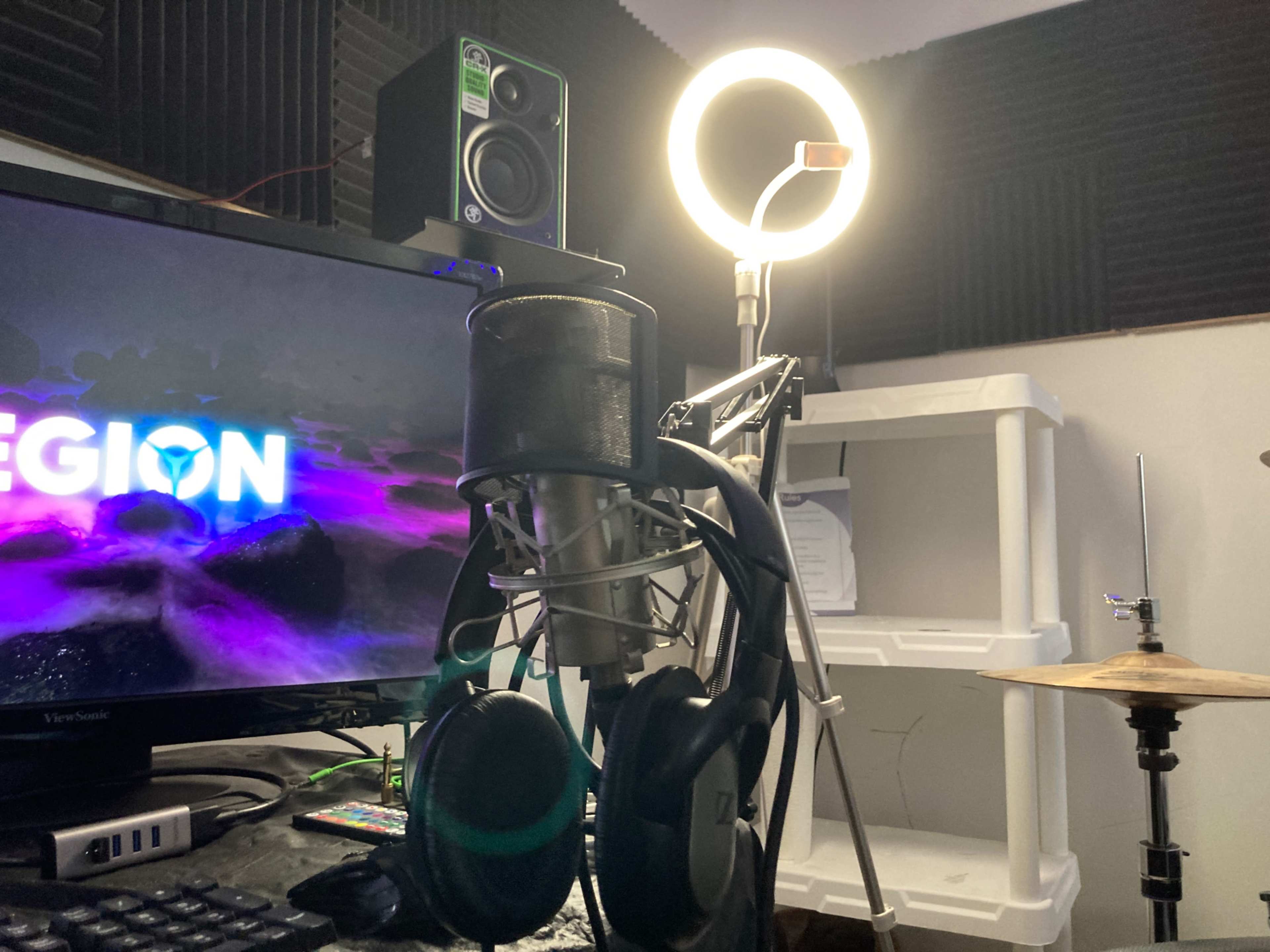 A microphone and headphones hang on a stand beside a computer monitor displaying the word "LEGION" and a ring light in a studio setup.