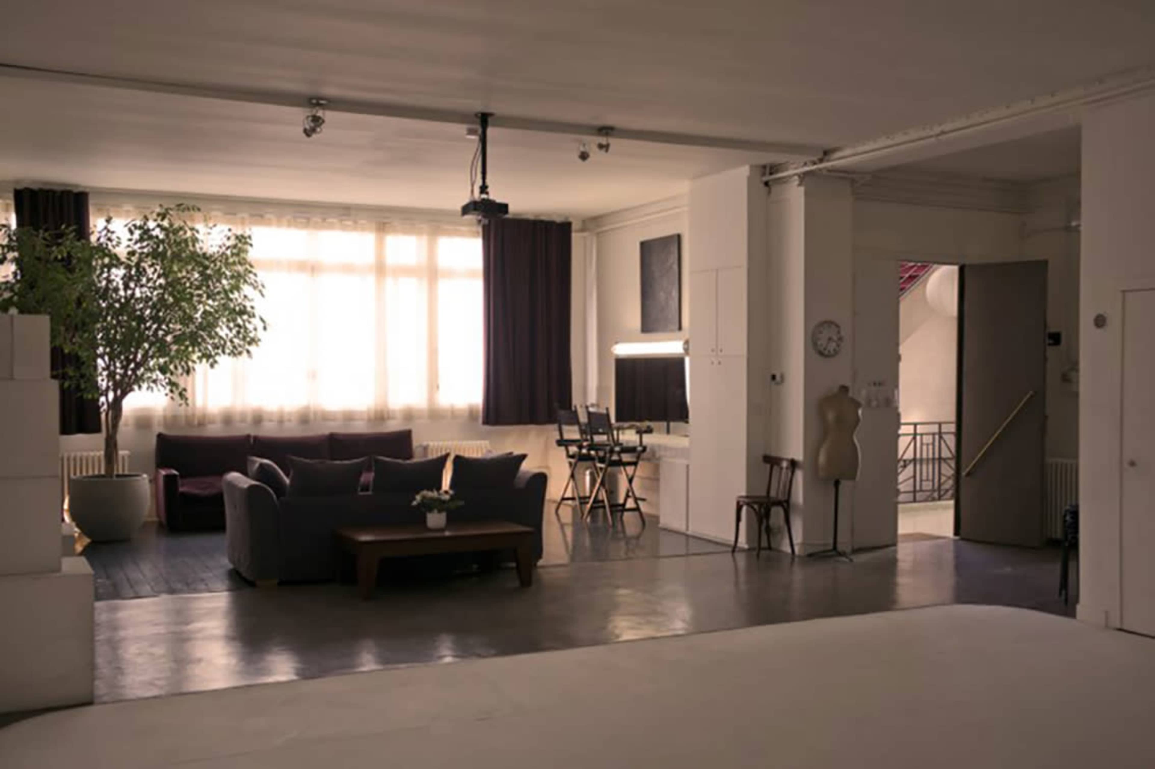 A spacious room with large windows, a dark sofa, a coffee table, a desk with a computer, and a doorway leading to another area.