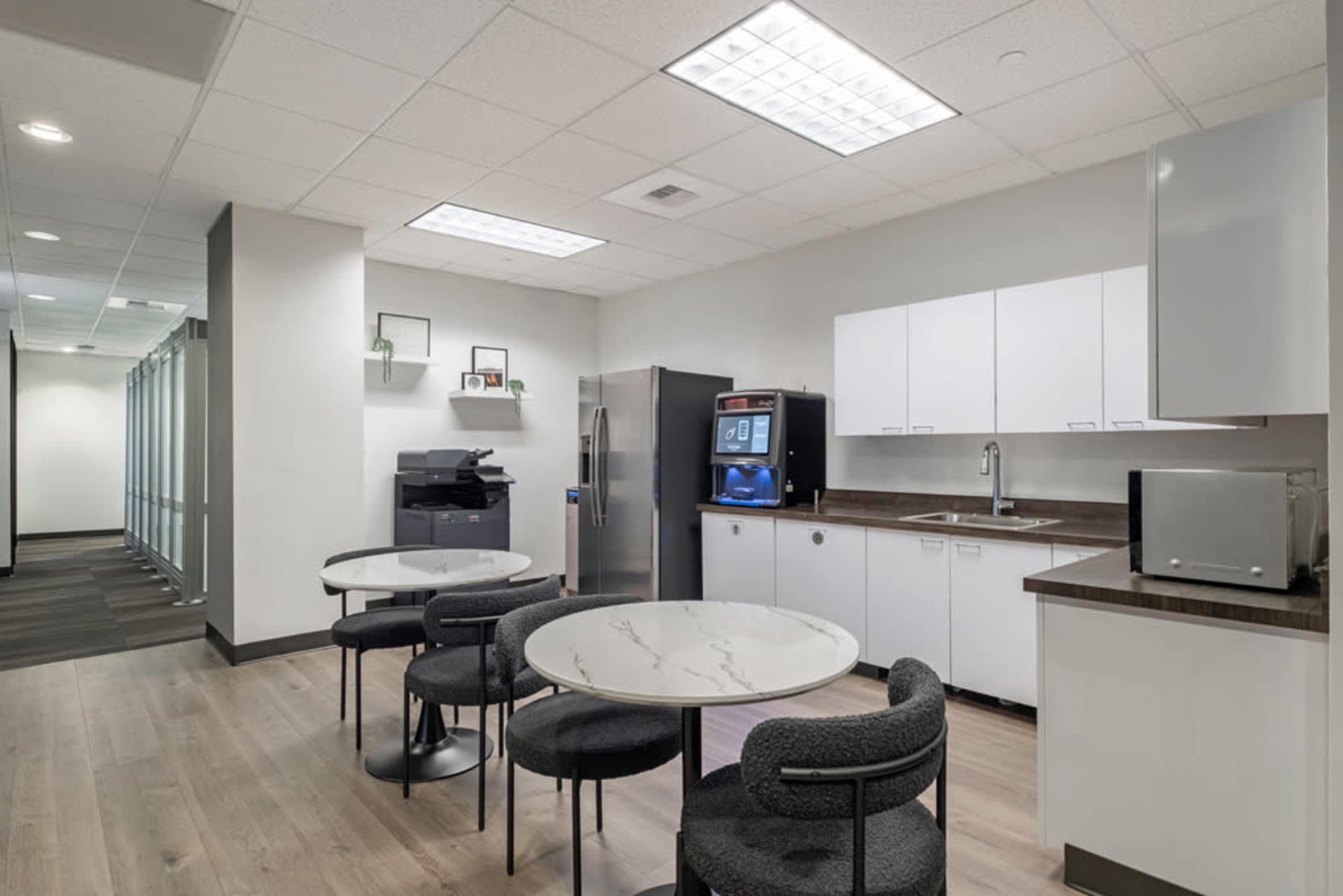 The image shows a modern office break room featuring a seating area with round tables, a refrigerator, a coffee machine, and cabinets along the wall.