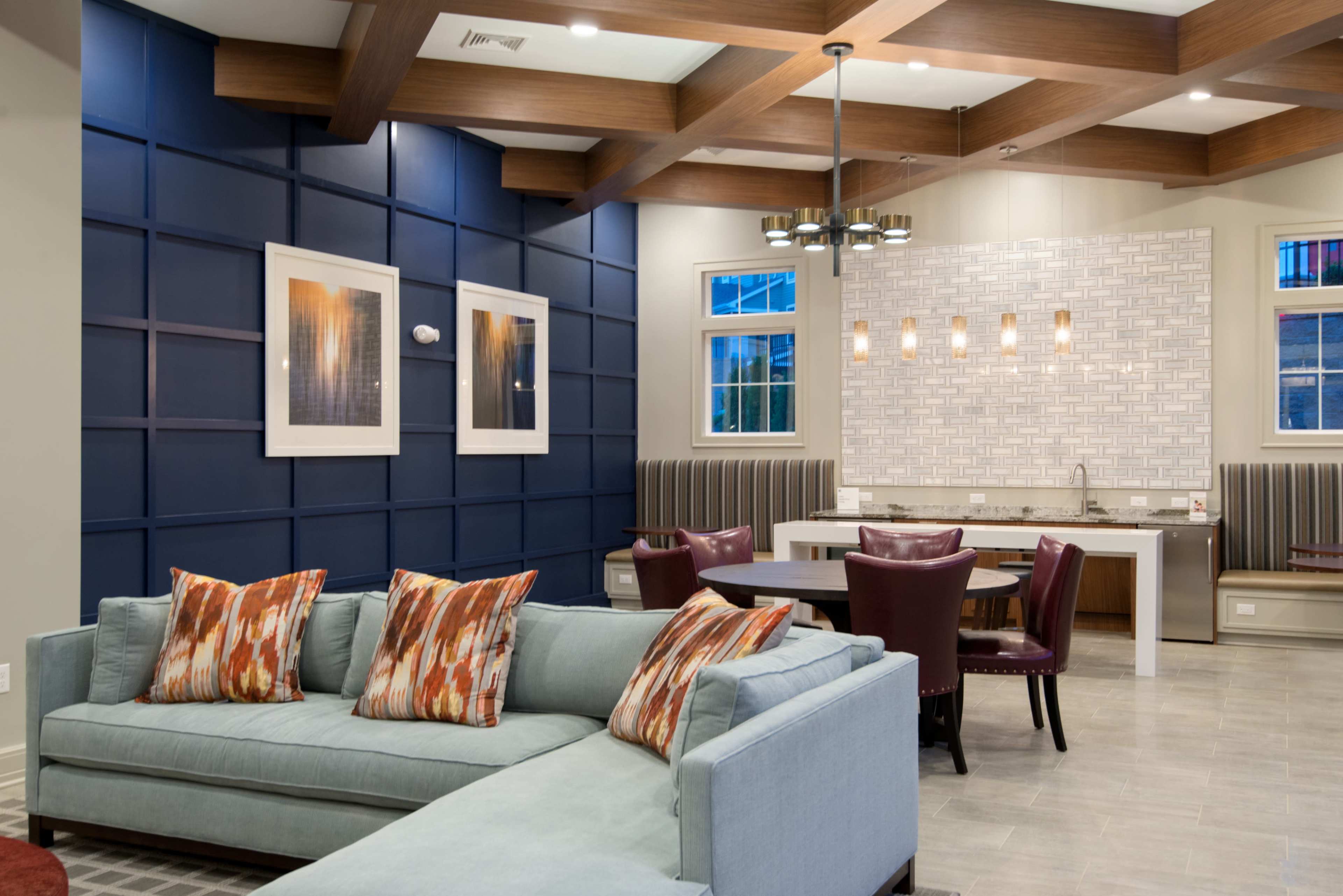 A modern lounge area featuring a light blue sectional sofa with decorative pillows, a dining area with a round table and burgundy chairs, and a kitchen space with white cabinetry and a tiled backsplash.