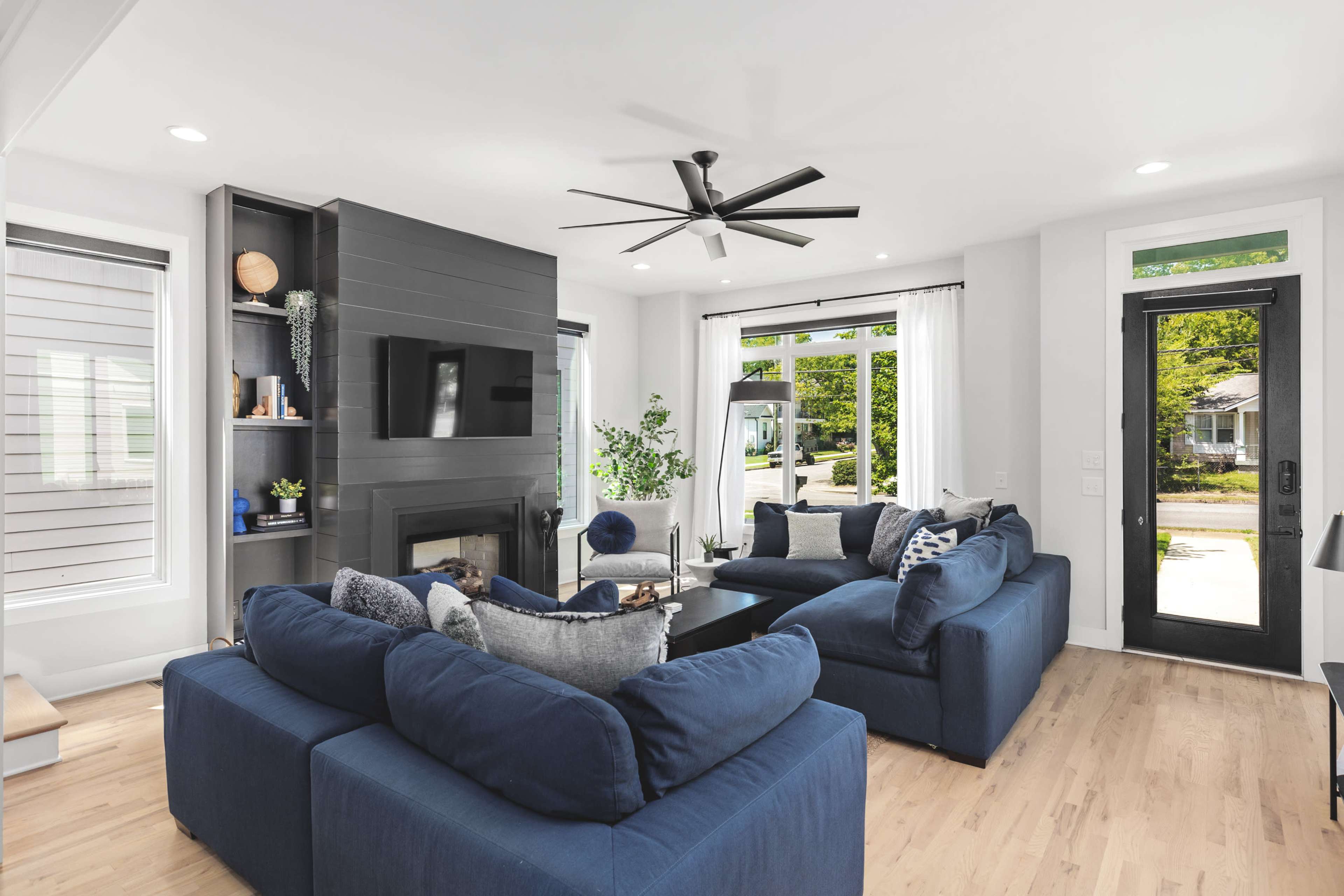 A modern living room features two blue couches arranged around a coffee table, with a fireplace and a large window providing natural light.