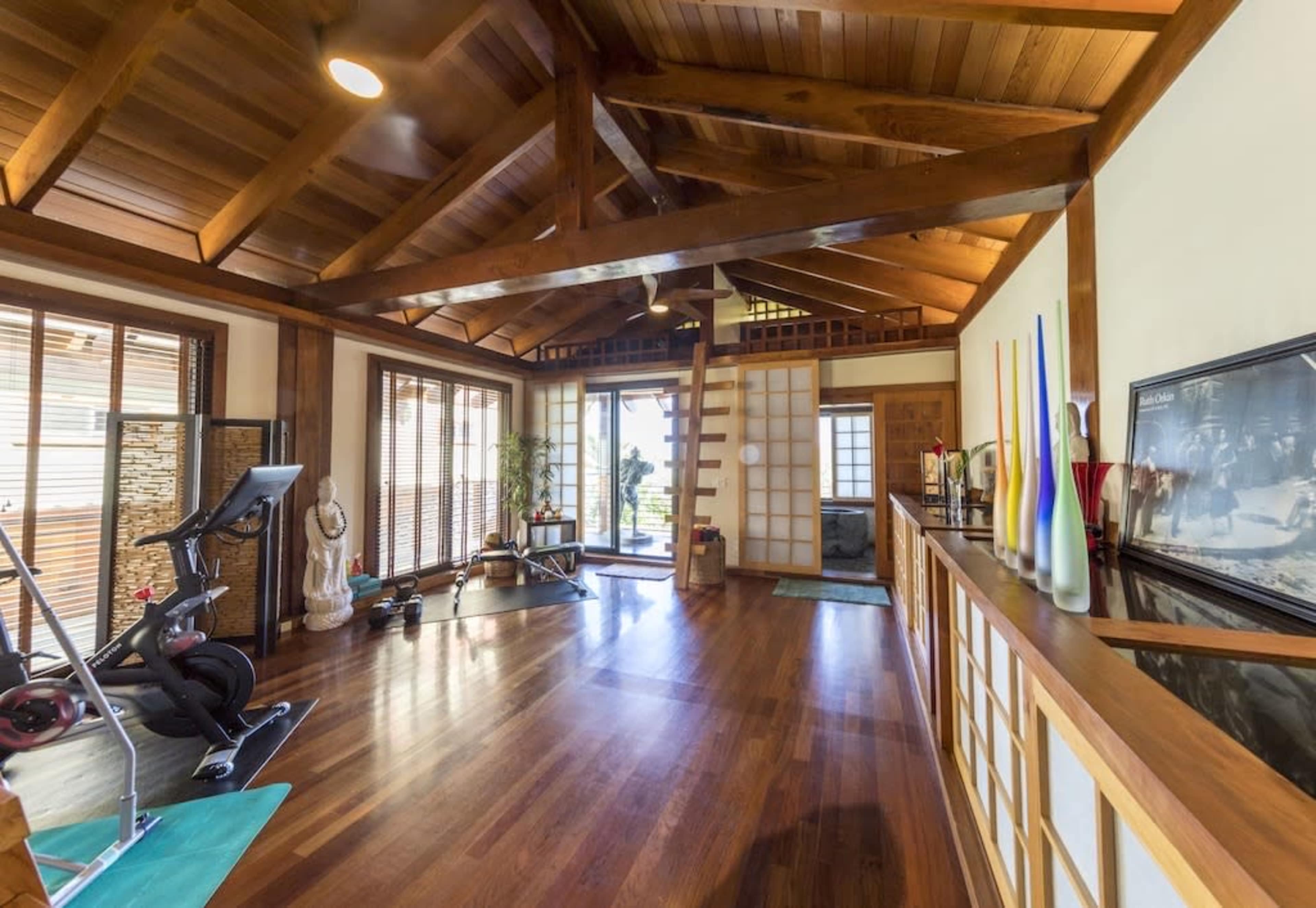 An interior of a spacious wooden room with a high ceiling, featuring exercise equipment, large windows, and a staircase leading to an upper level.