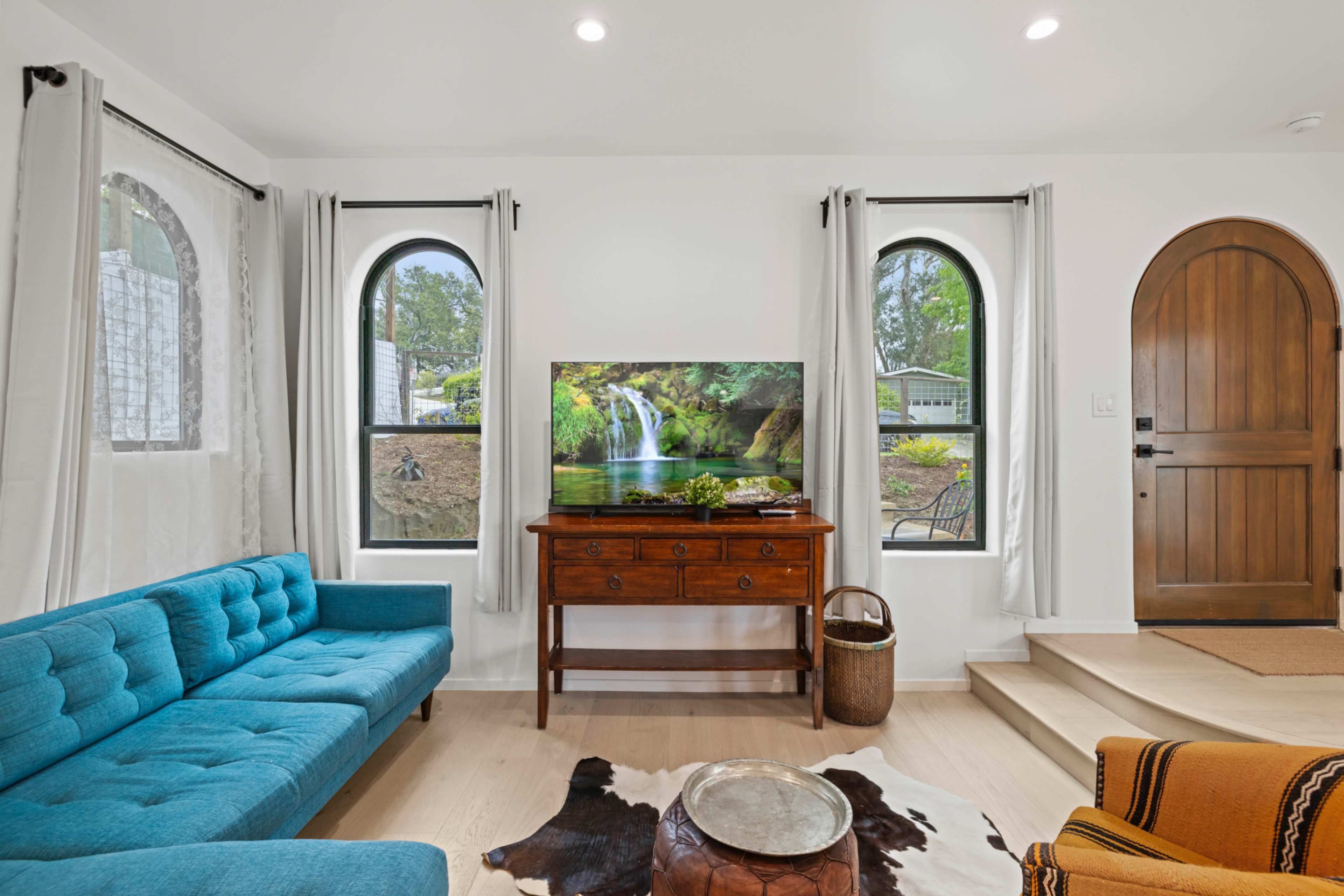 The living room features a blue sofa, a wooden chest, a large television displaying a nature scene, and an arched doorway leading to the outside.
