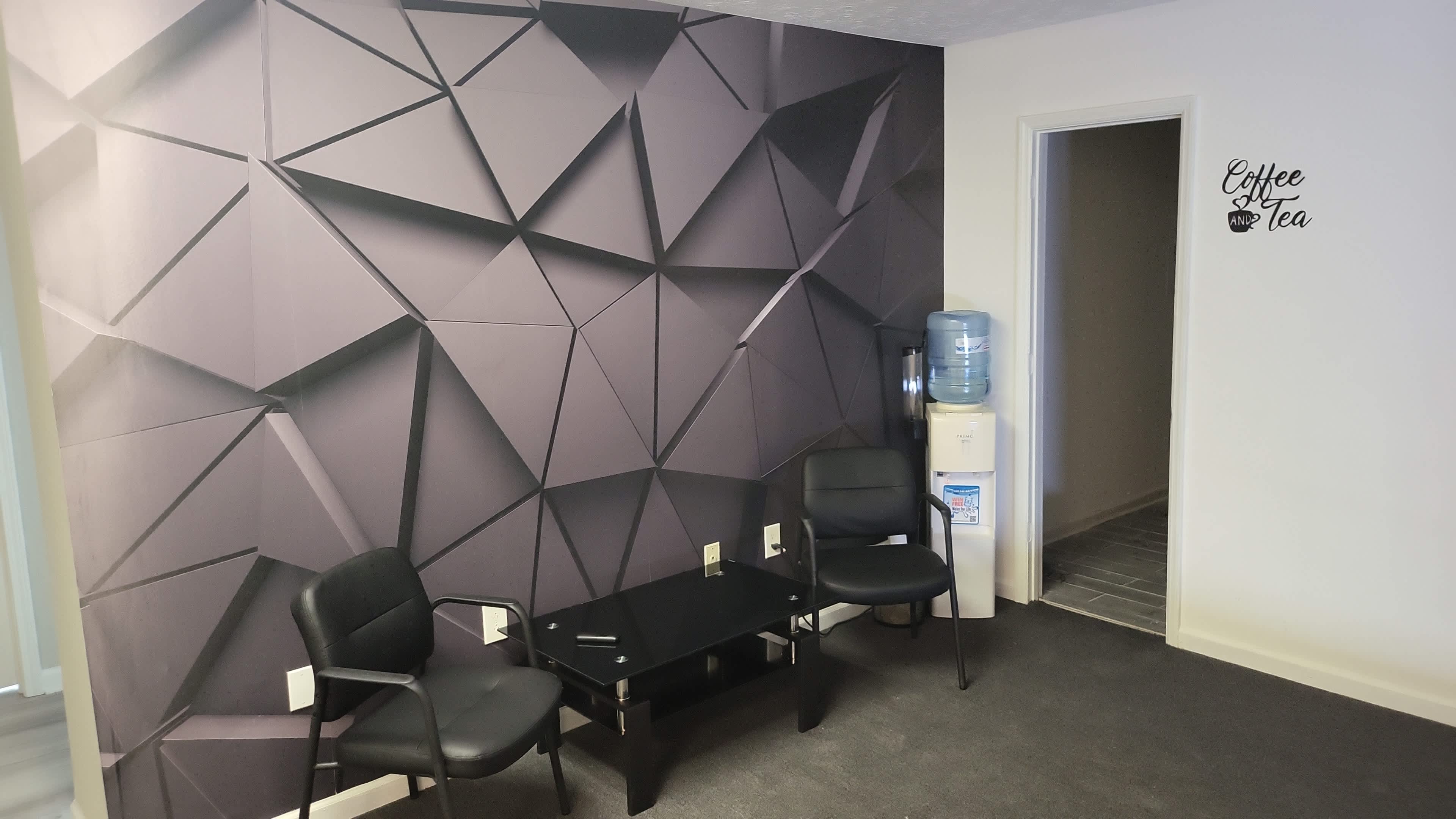 The image shows a small waiting area with two black chairs, a glass table, a geometric patterned wall, and a water cooler near an entrance.