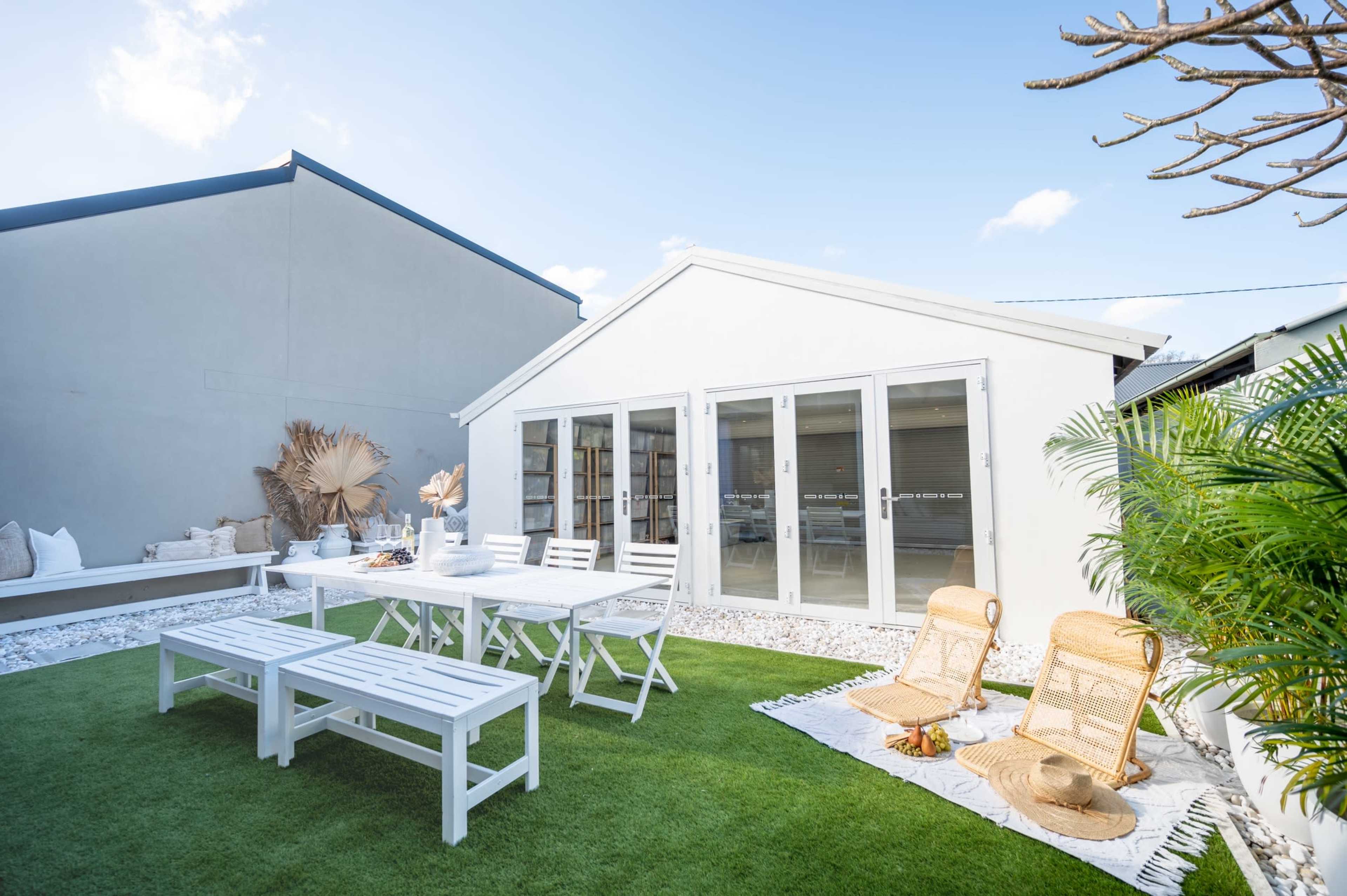 A white garden shed with large glass doors is surrounded by a green lawn, outdoor furniture, and decorative plants.