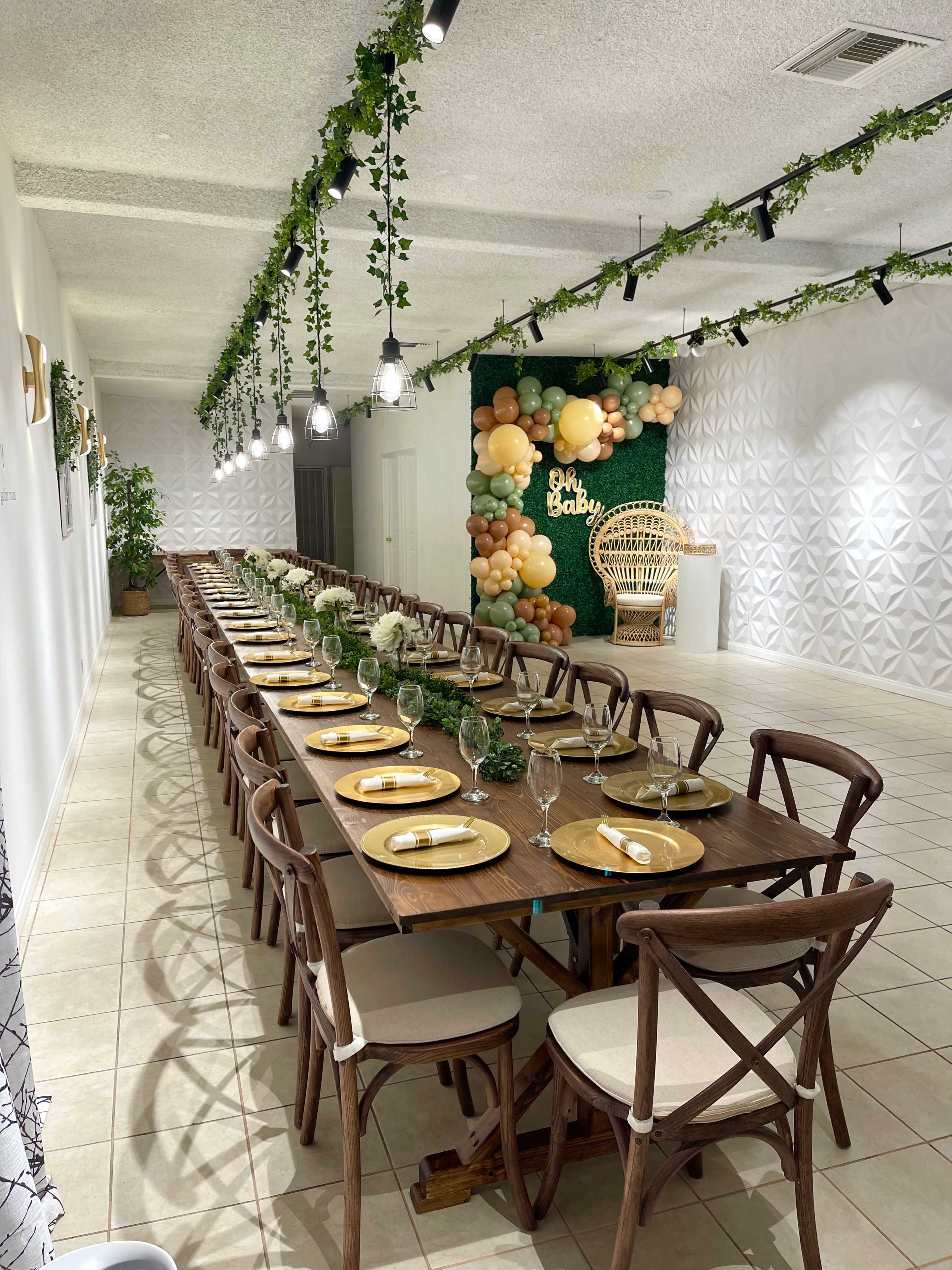 The image shows a long, elegantly arranged dining table with wooden chairs set for a special event, adorned with greenery and decorative balloons in a well-lit room.