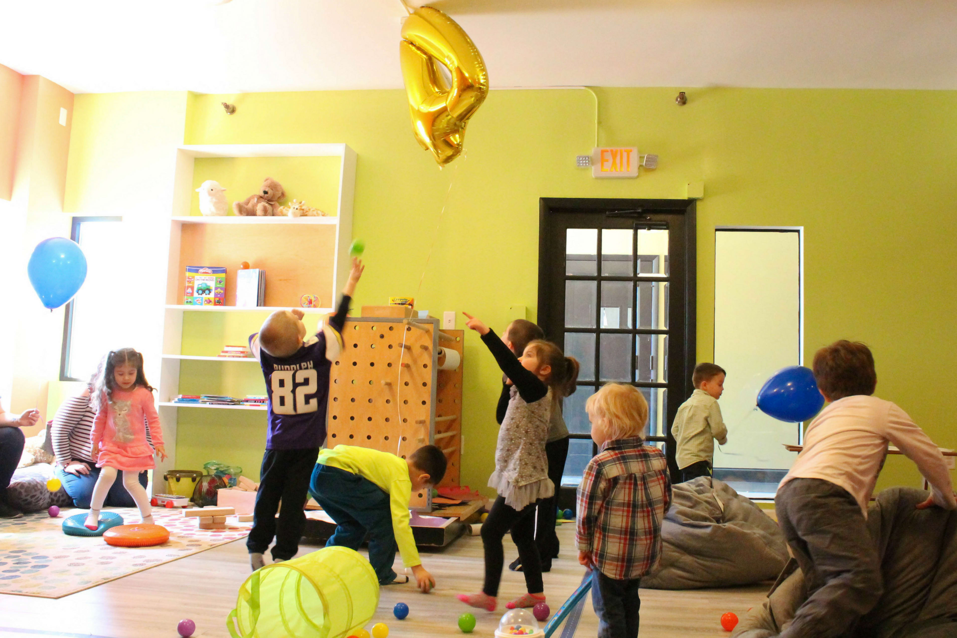A group of children play with colorful toys and balloons in a brightly lit room with shelves and a carpeted area.