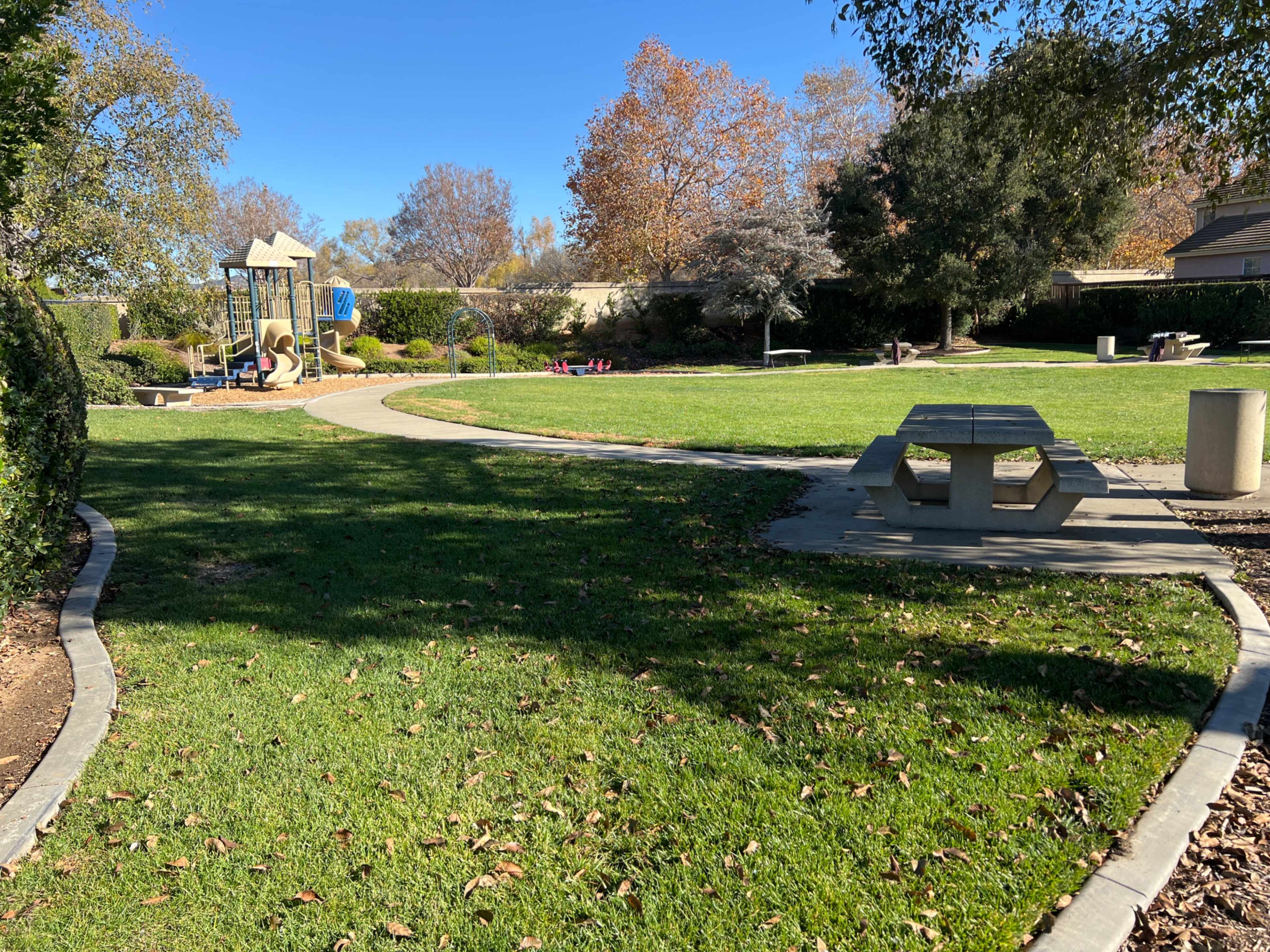 A park features a path leading to a playground, surrounded by grass and trees, with a picnic table in the foreground.