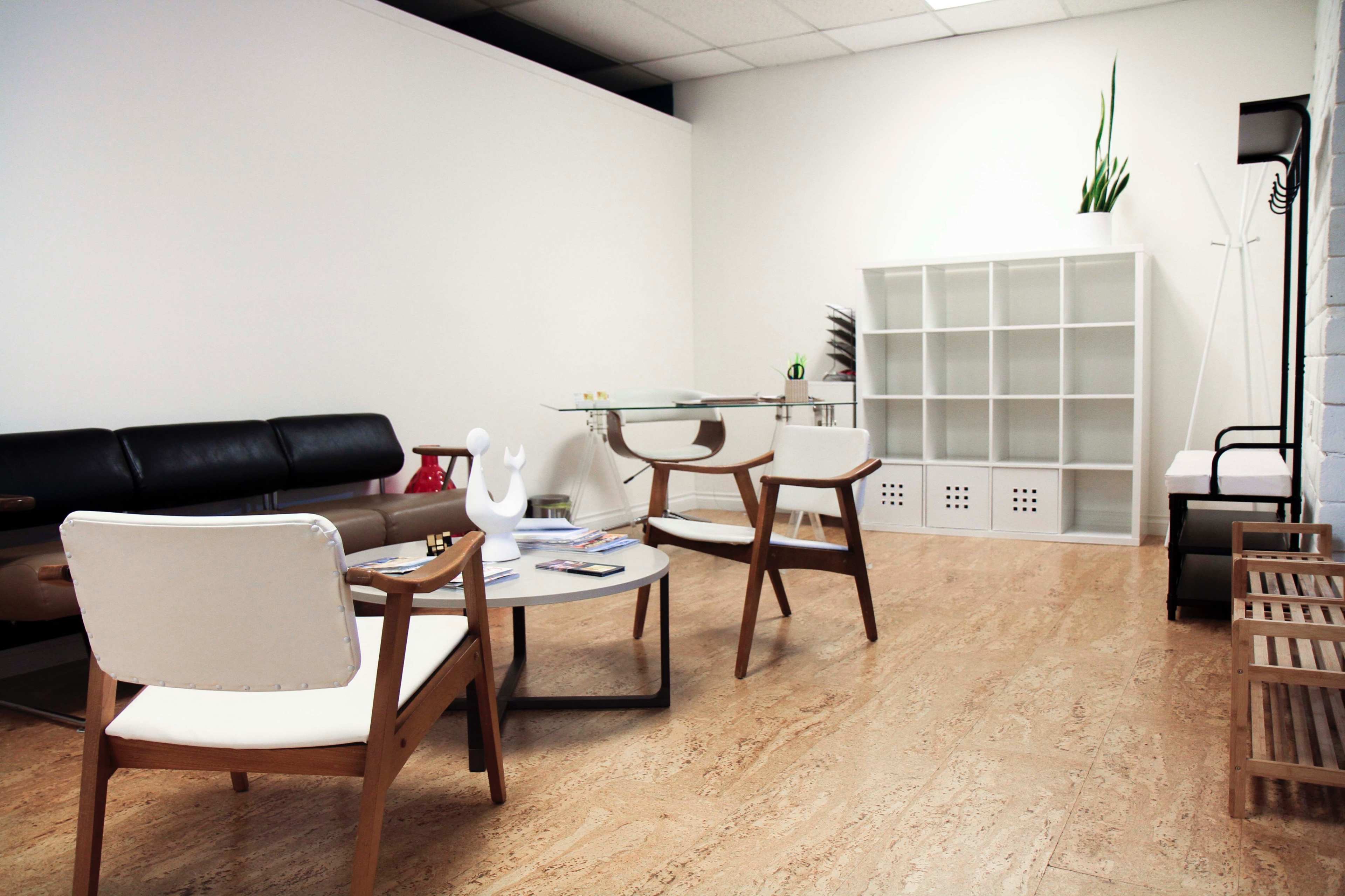 A minimalist office waiting area with a black leather sofa, two wooden chairs, a glass coffee table, and a white shelving unit against a plain wall.