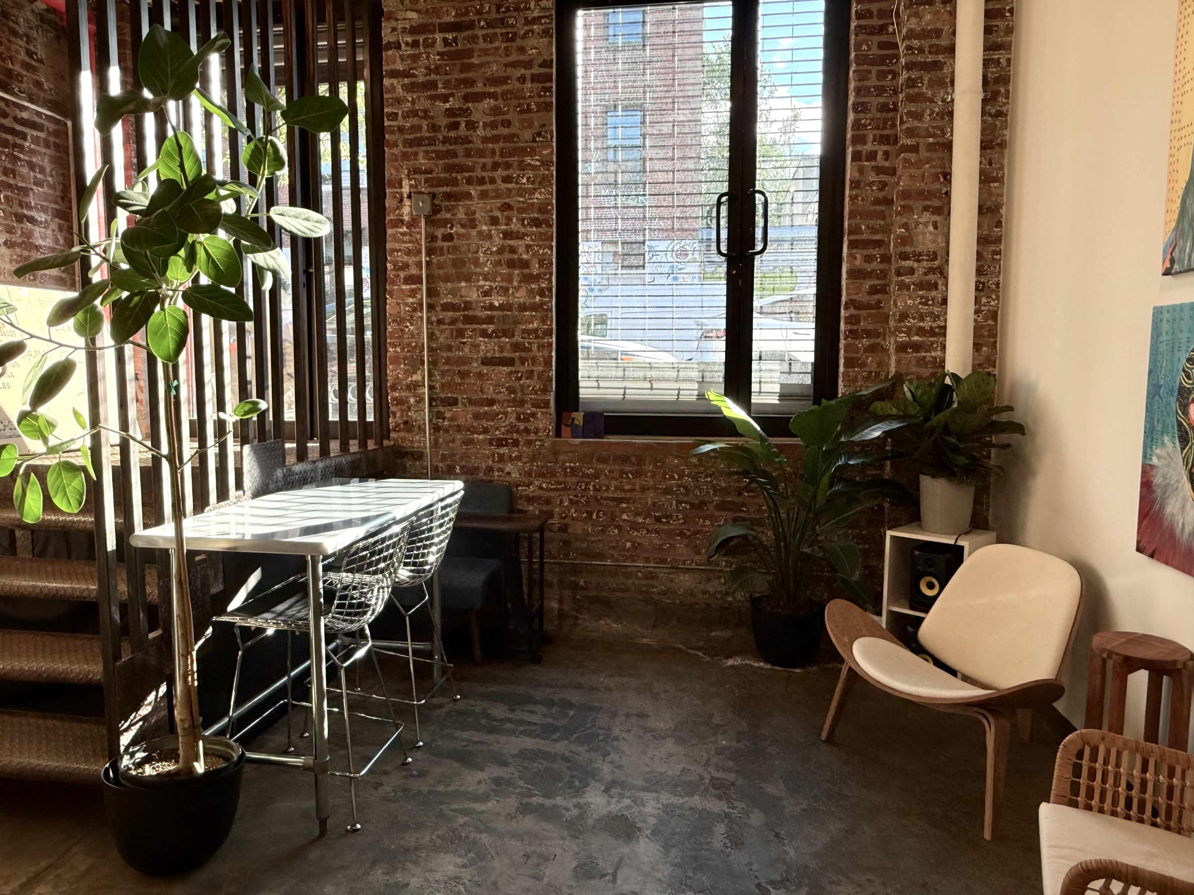 The image shows a cozy interior space with exposed brick walls, a plant near a glass table and stools, and a comfortable chair next to a window.