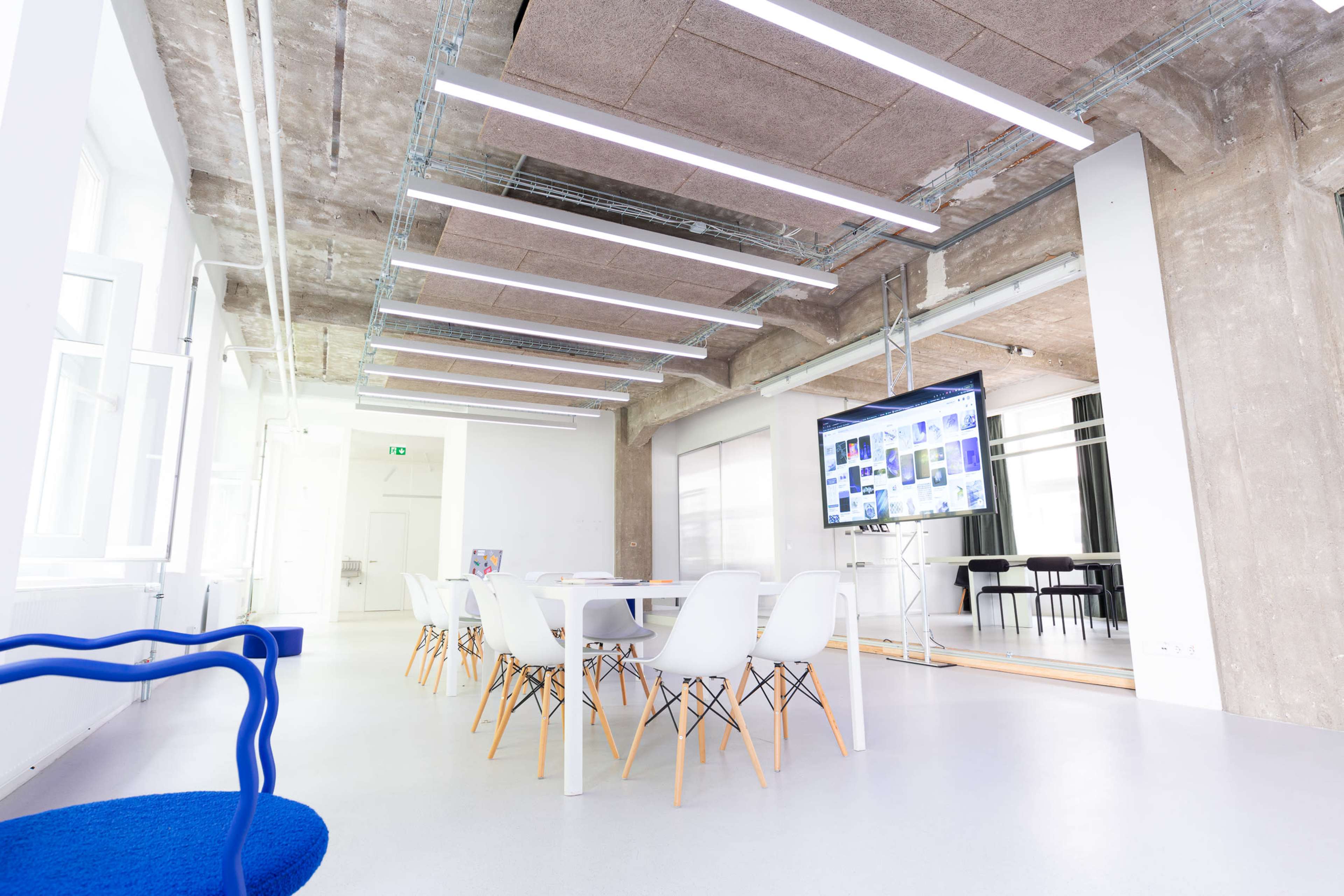 A modern conference room features a long white table surrounded by chairs, with exposed concrete walls and bright overhead lighting.