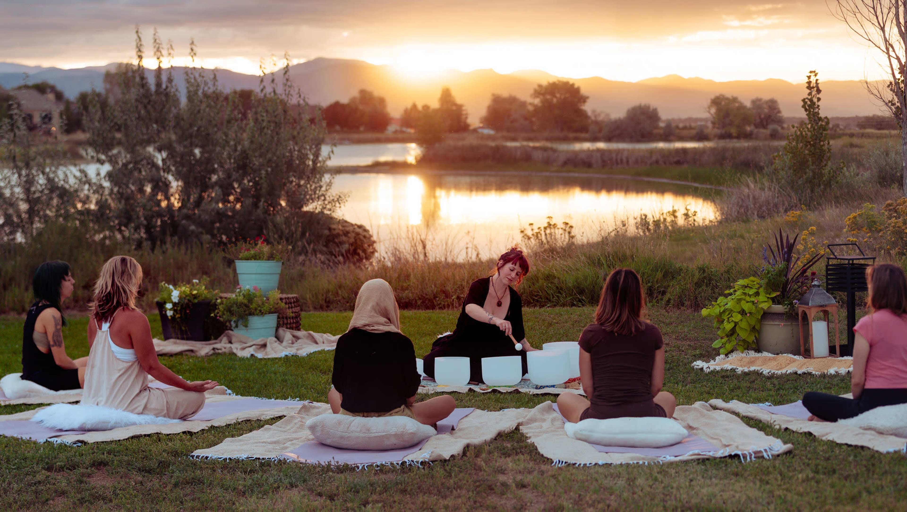The scene depicts a group of people seated on mats in a grassy area by a lake at sunset, with one individual playing crystal bowls.