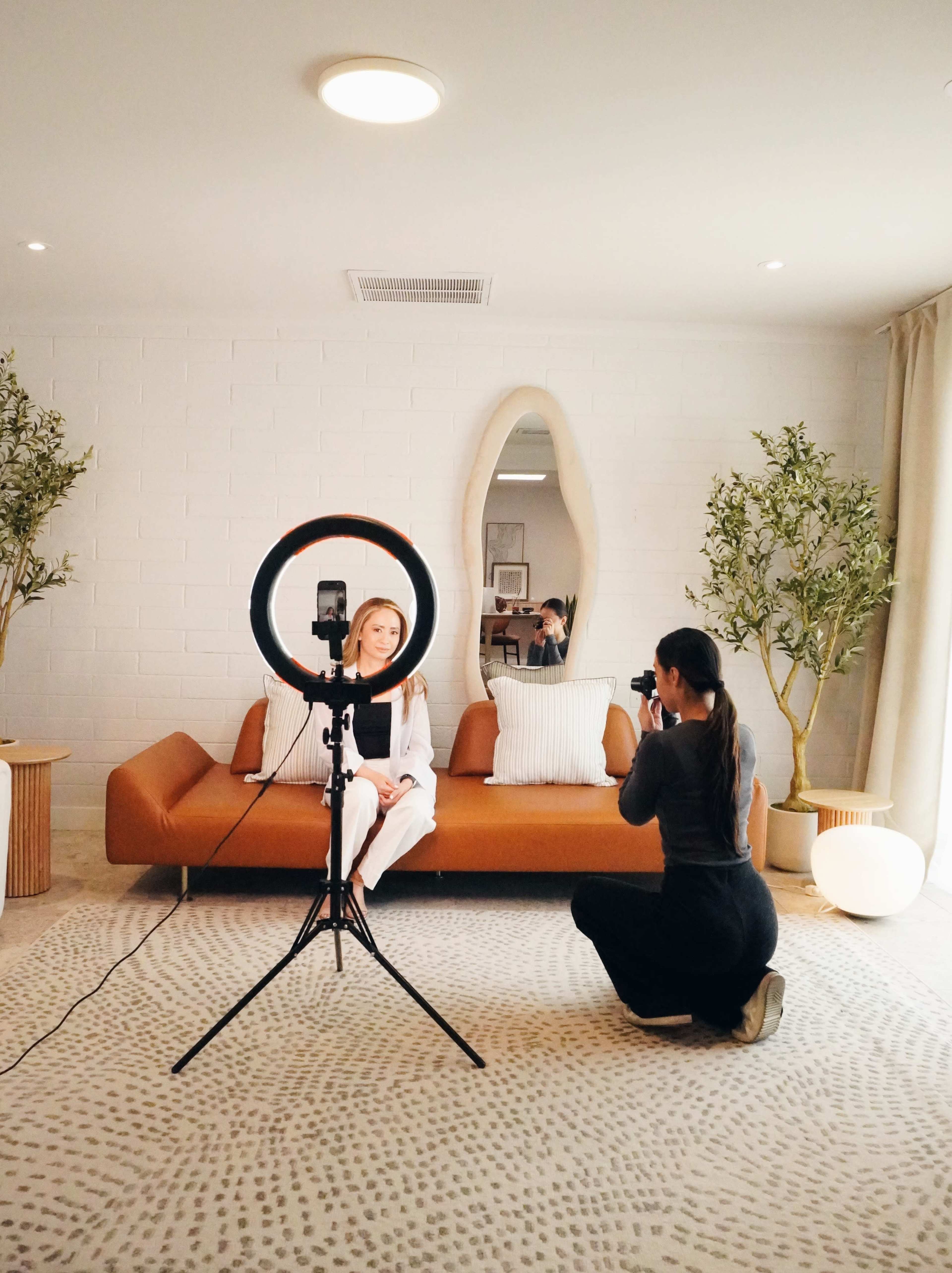 A woman sits on a brown sofa as another woman photographs her using a ring light in a well-lit room.