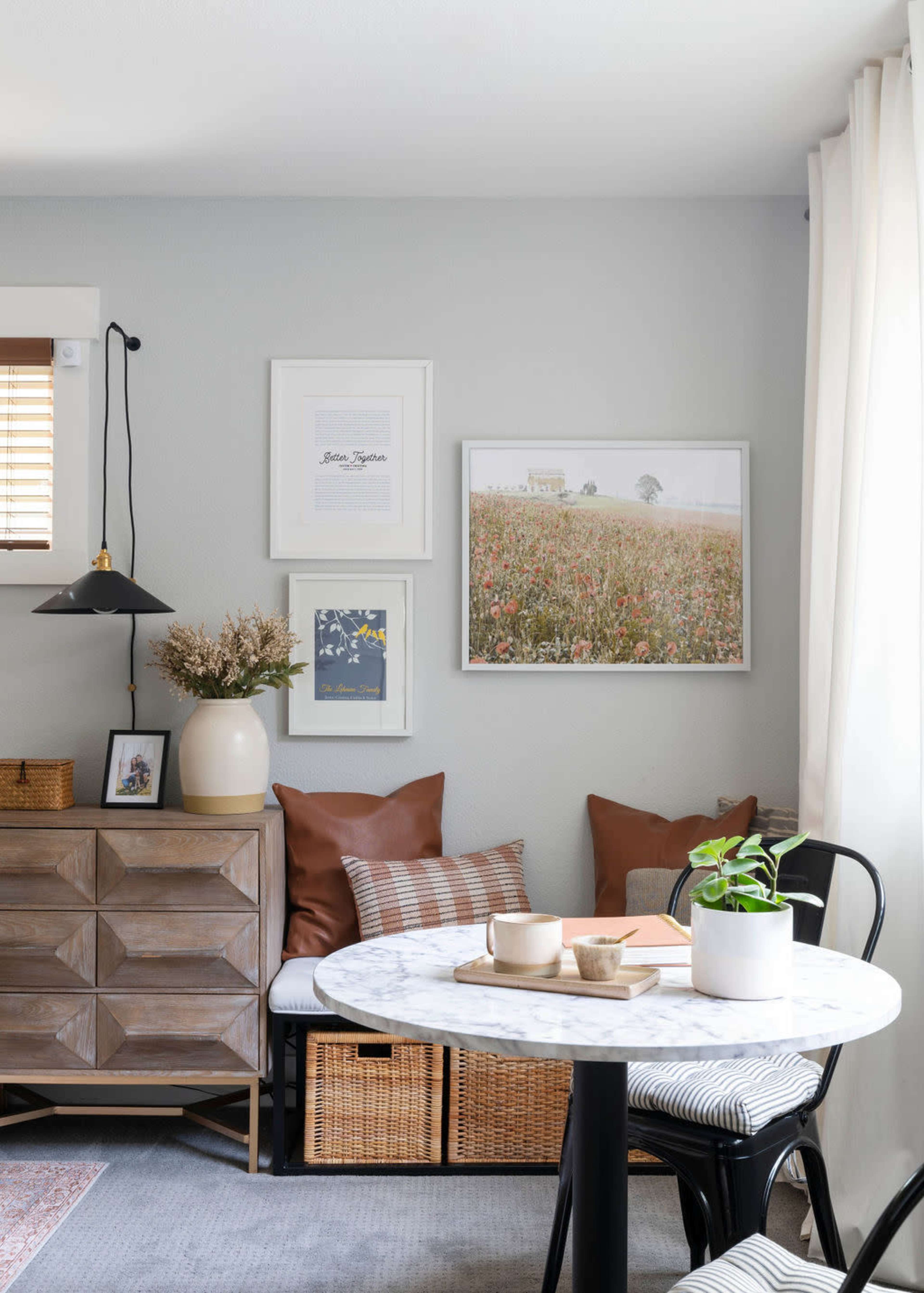 A small dining area features a round marble table, a wooden sideboard, and framed artwork on the gray walls, with cushions on a built-in bench and a potted plant on the table.