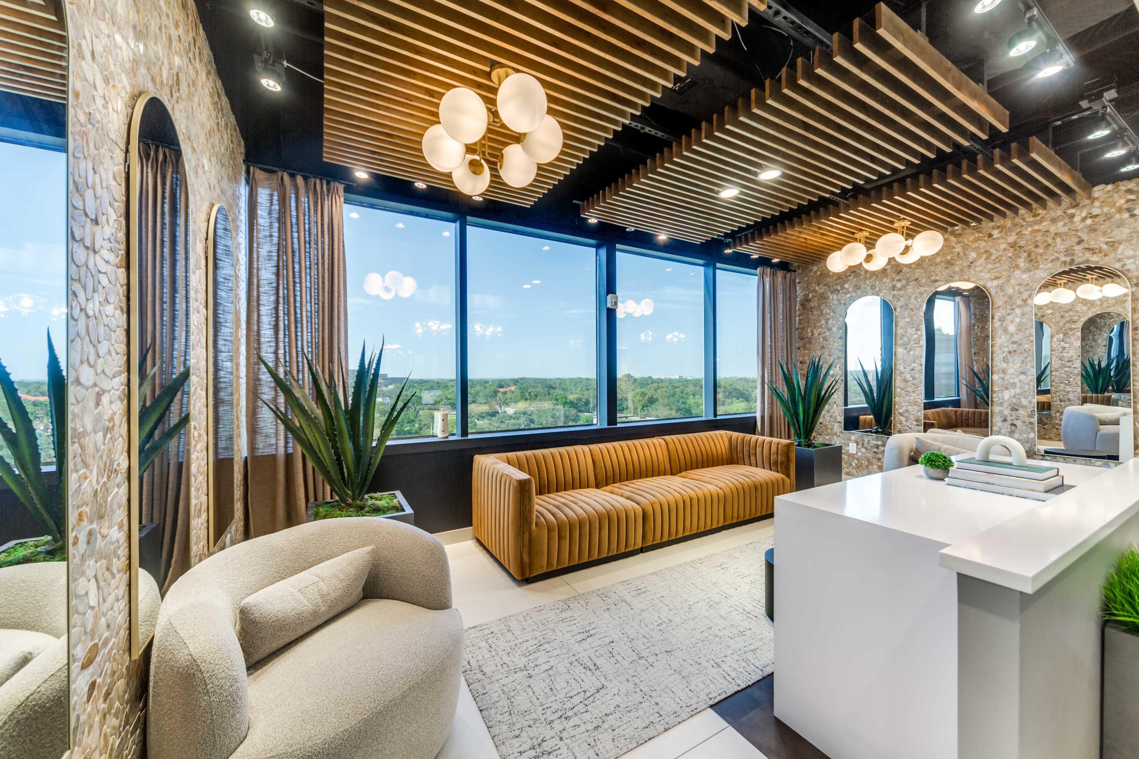 The image shows a modern lounge area with a curved sofa and a striped couch, surrounded by large windows offering a view of green landscape, and features wooden ceiling panels and decorative plants.