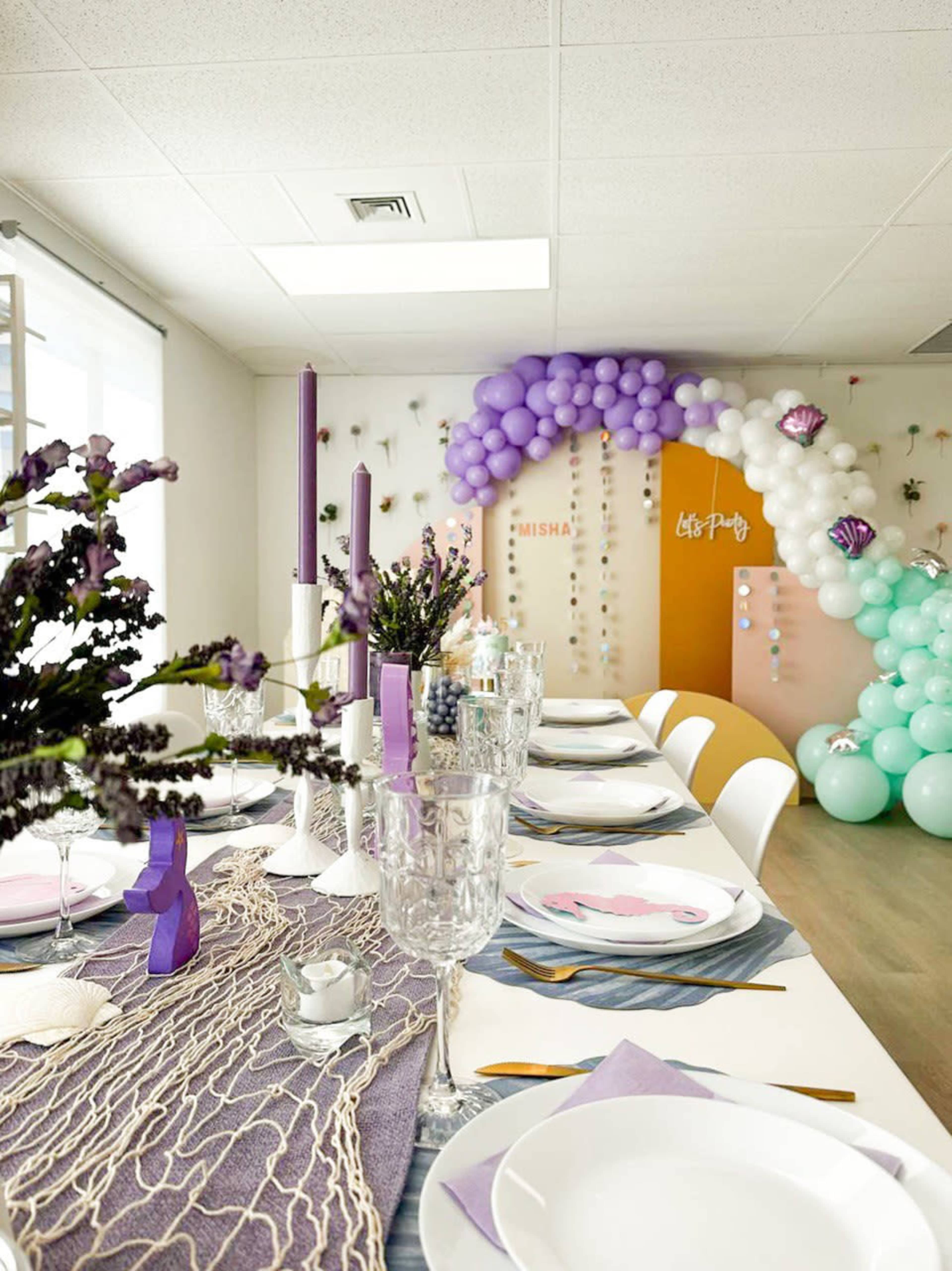 A party setup features a decorated table with lavender accents, crystal glassware, and a colorful balloon arch in the background.
