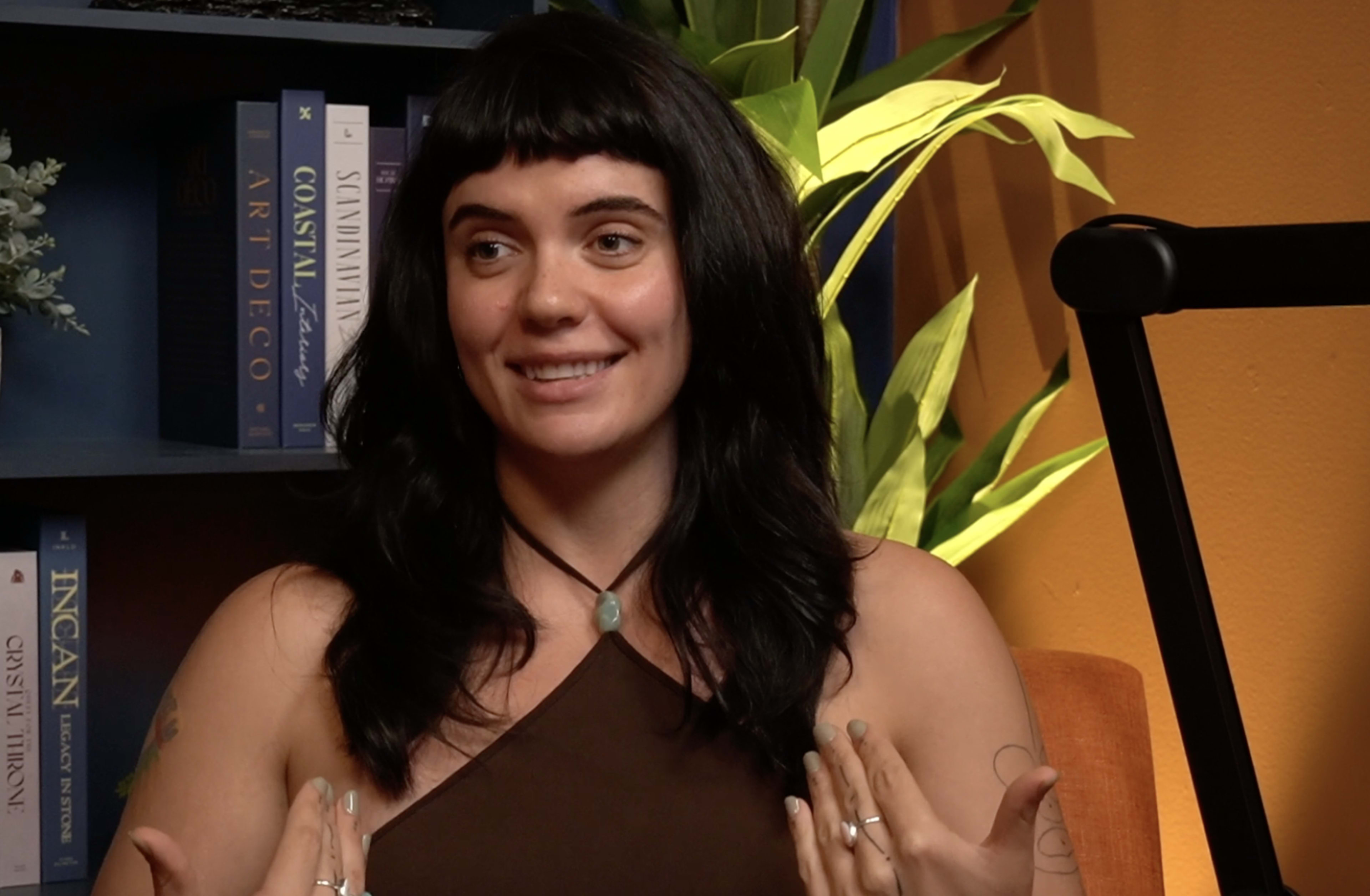 A person with long black hair is sitting on a couch and speaking in front of a bookshelf filled with books and a decorative plant.