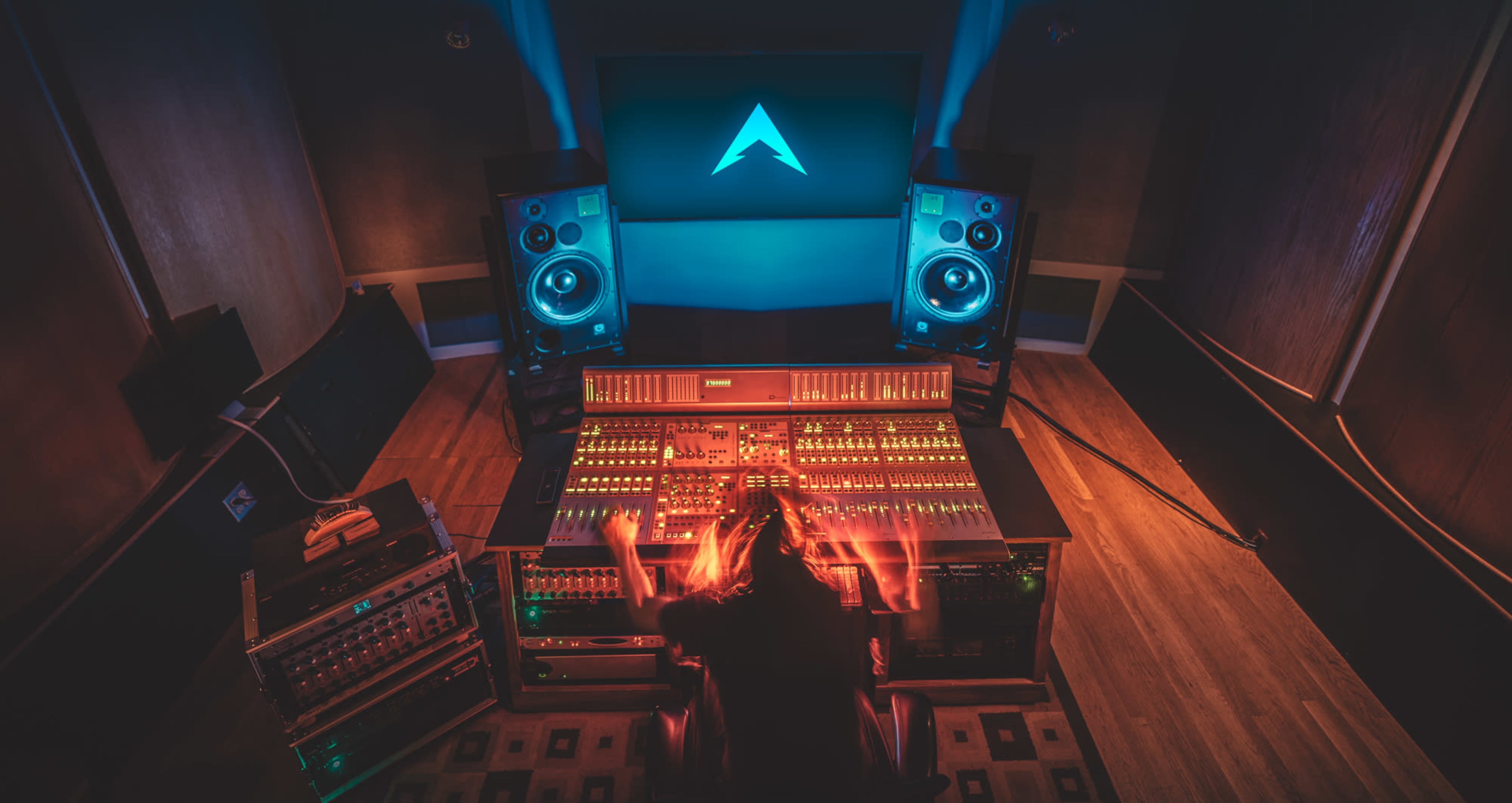 A person is working at a large audio mixing console in a sound studio, surrounded by loudspeakers and a large screen displaying a logo.