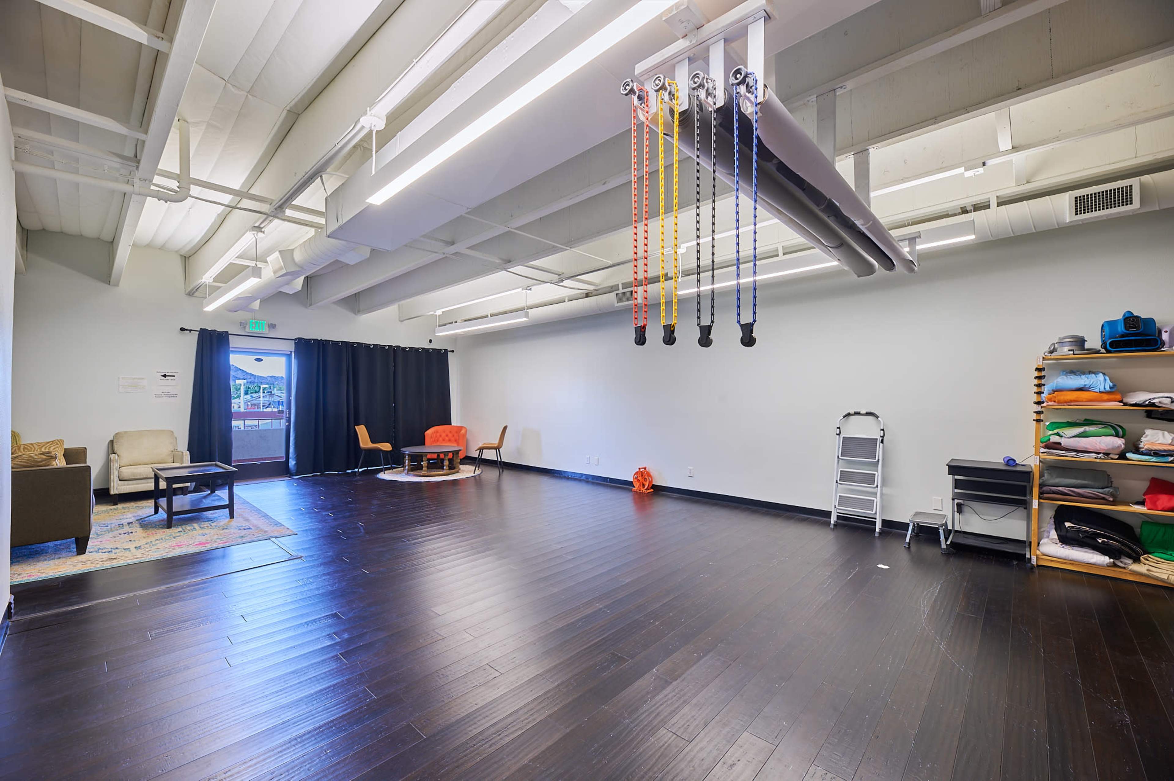 The image shows an empty exercise studio with wooden flooring, ceiling lights, and a few pieces of furniture along one side.