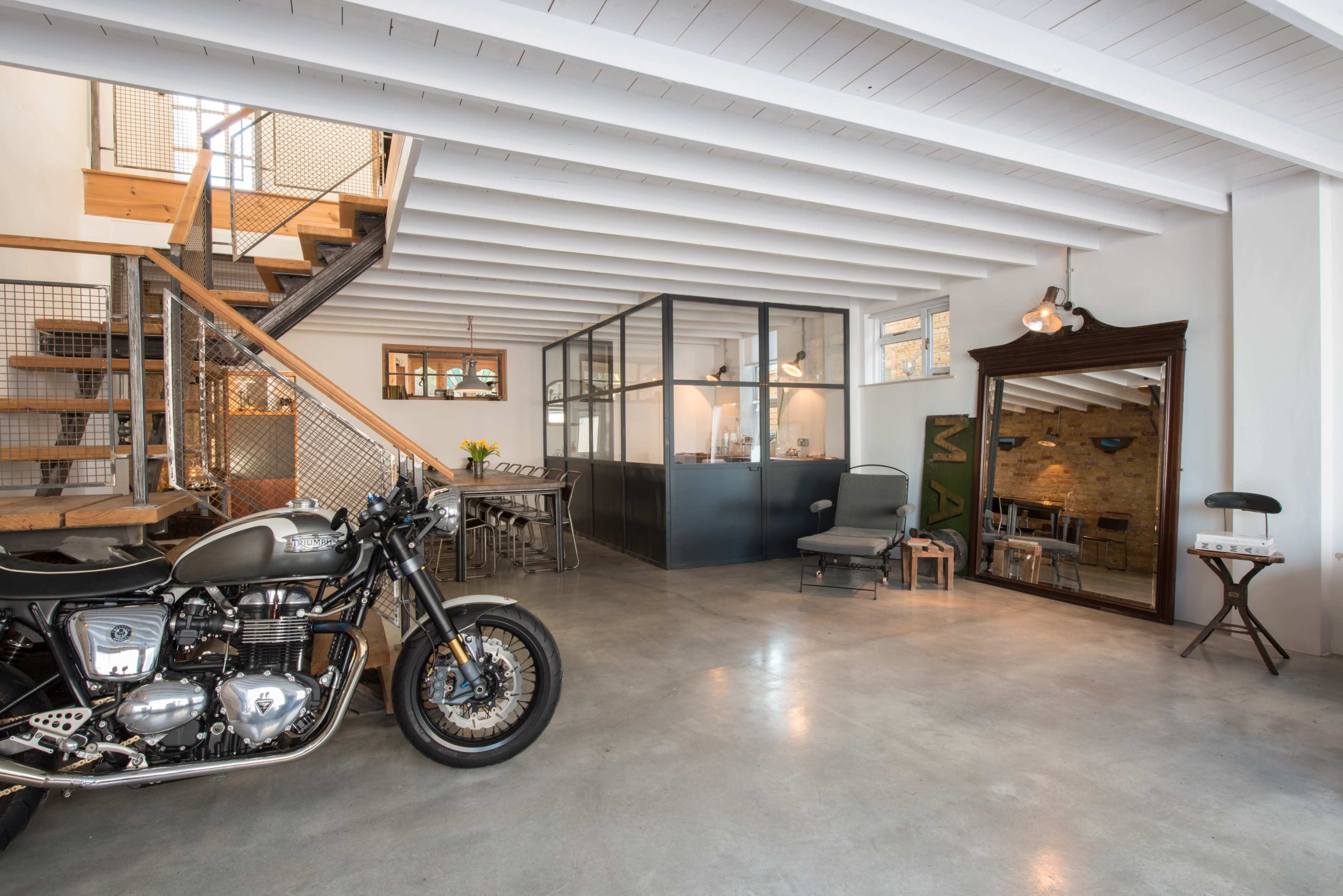A modern industrial-style interior features a motorcycle, a wooden staircase, and a glass-walled kitchen area.