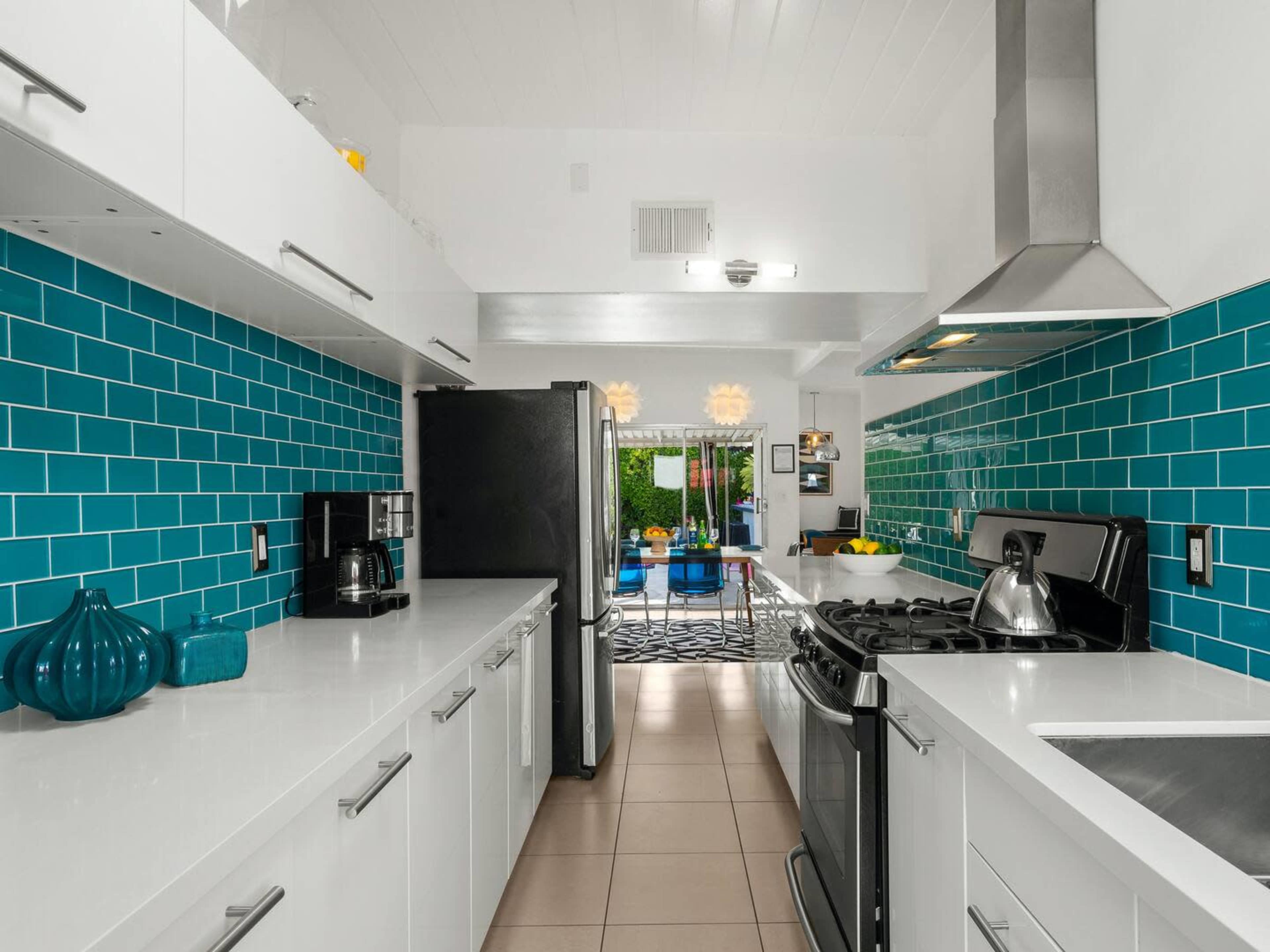A modern kitchen features turquoise tiled walls, white cabinetry, and stainless steel appliances, with a view of an outdoor patio through an open door.