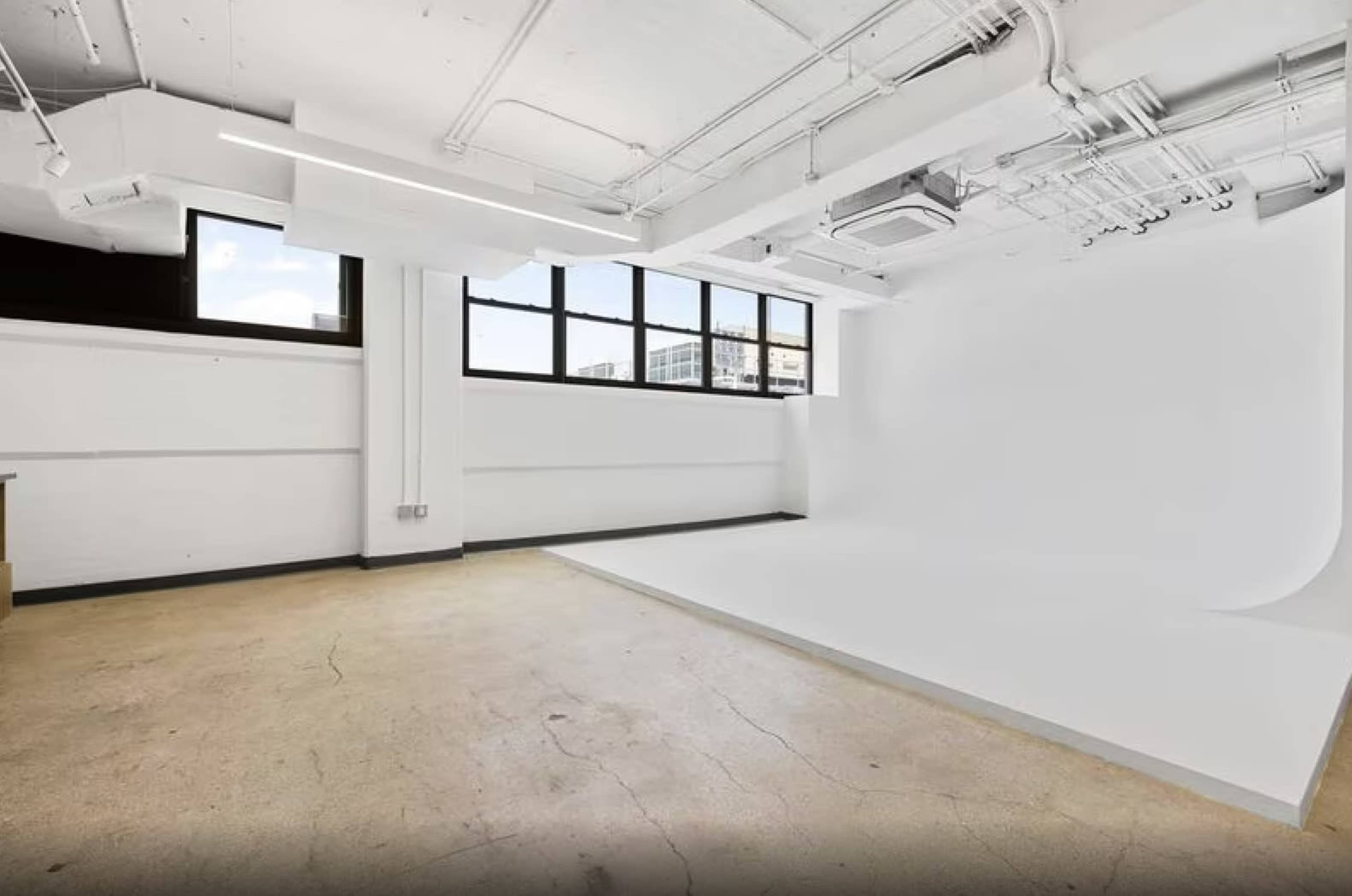 The image shows a spacious, empty studio with a white backdrop and large windows adjacent to a concrete floor.
