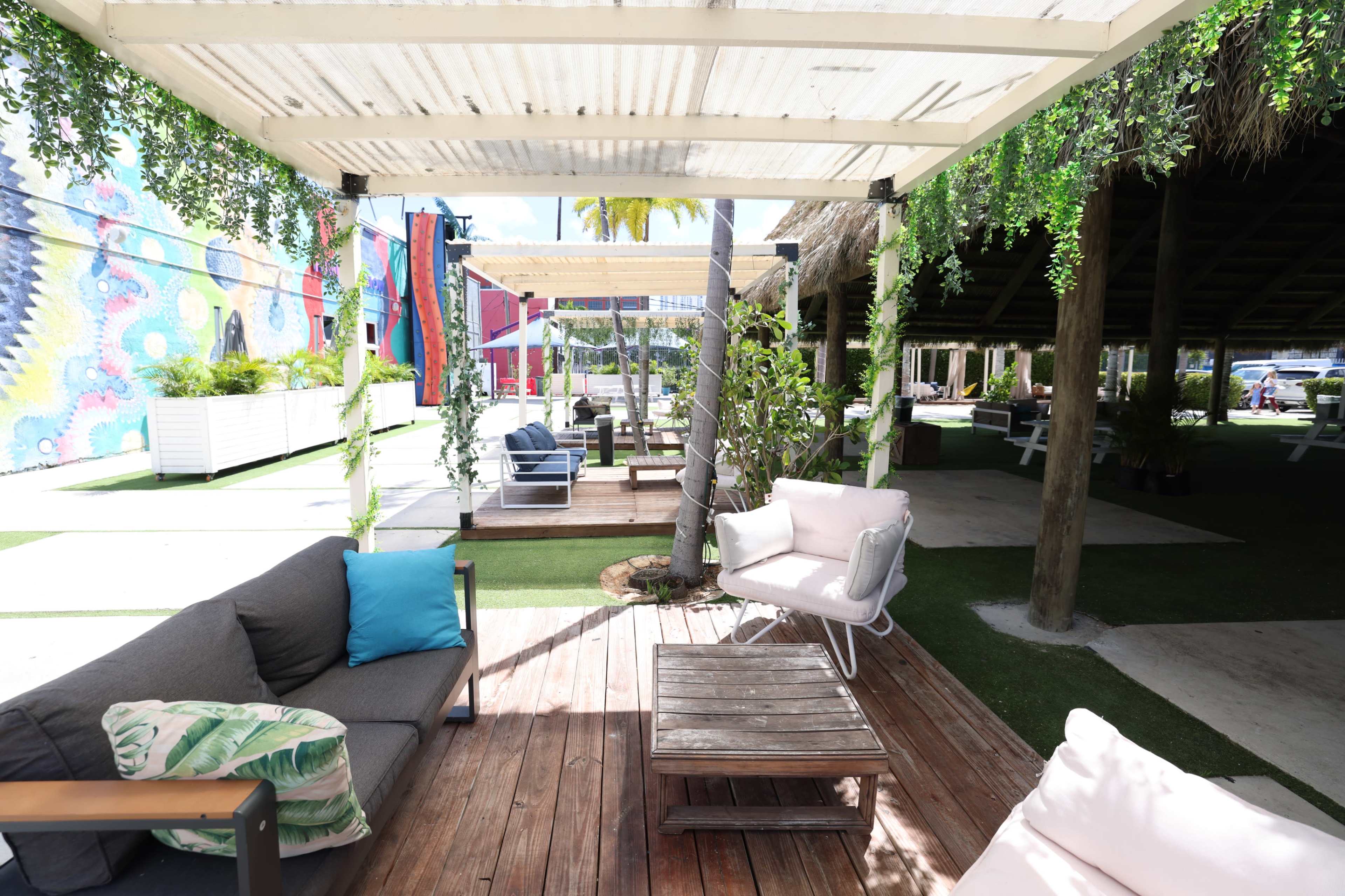 The image shows a shaded outdoor lounge area with wooden furniture, green plants, and vibrant murals in the background.