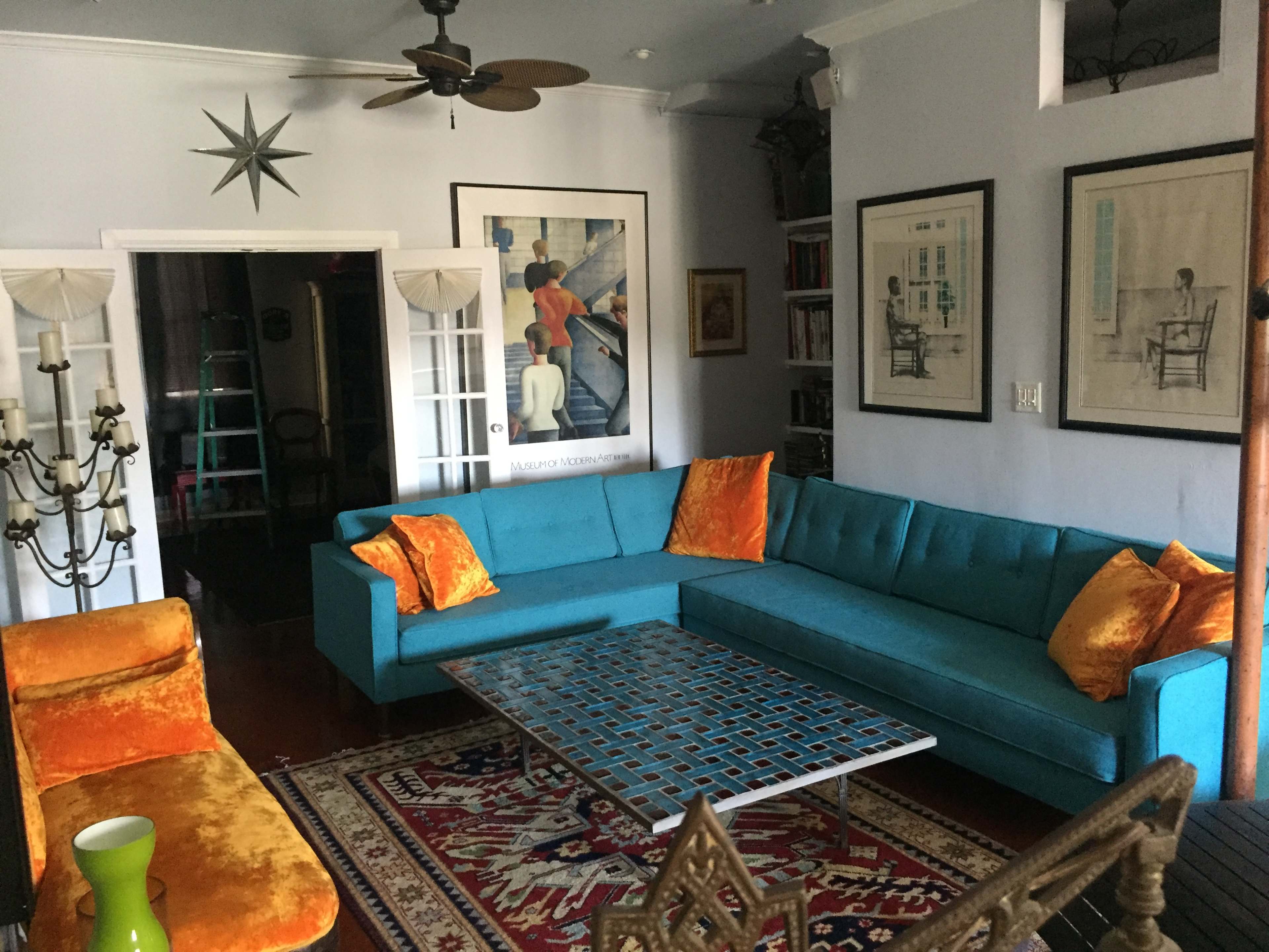 The room features a teal sectional sofa with orange throw pillows, a patterned coffee table, and framed artwork on the walls.