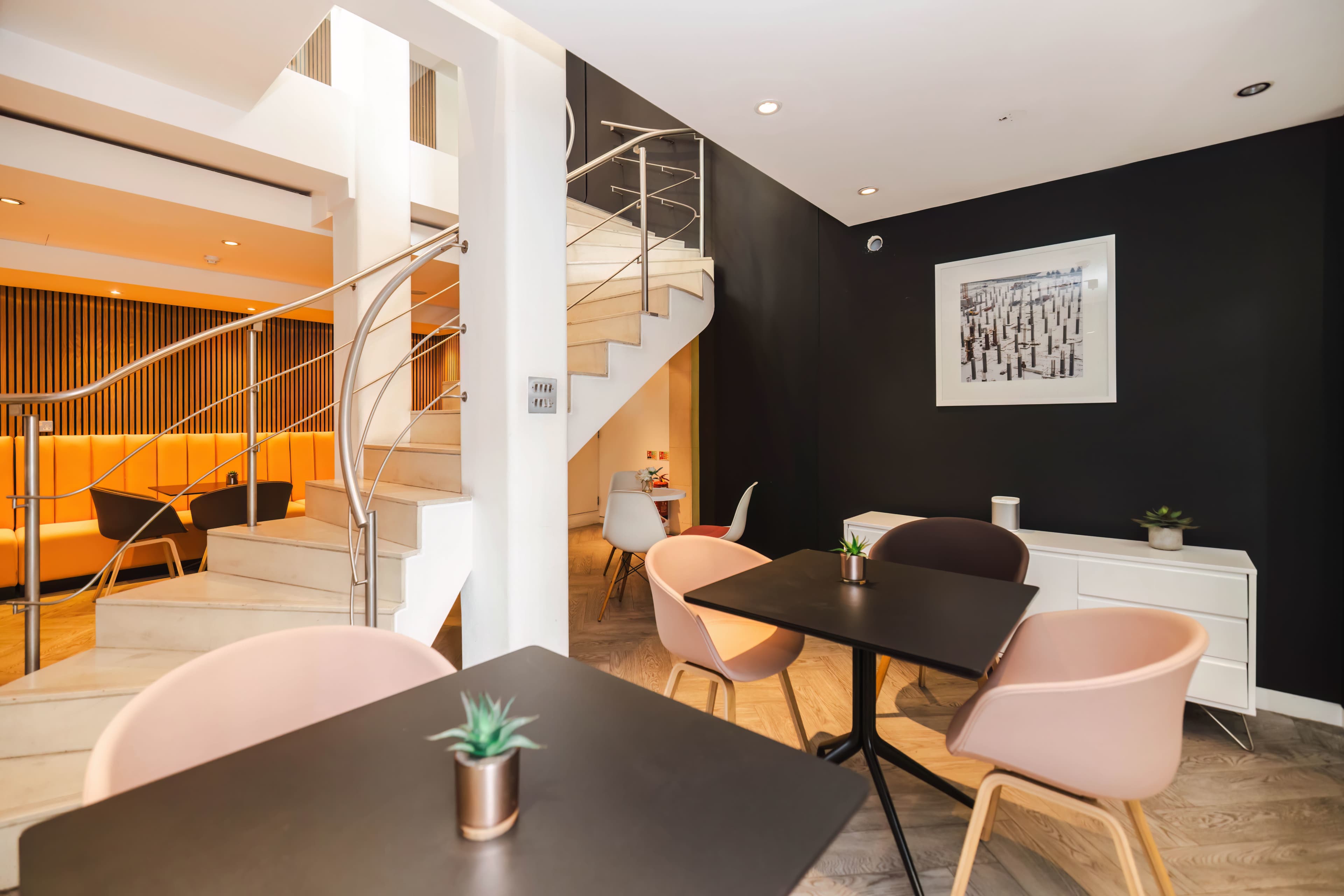 A modern café interior features white walls, wooden floors, and minimalist furniture, including tables and chairs arranged around a staircase leading to an upper level.