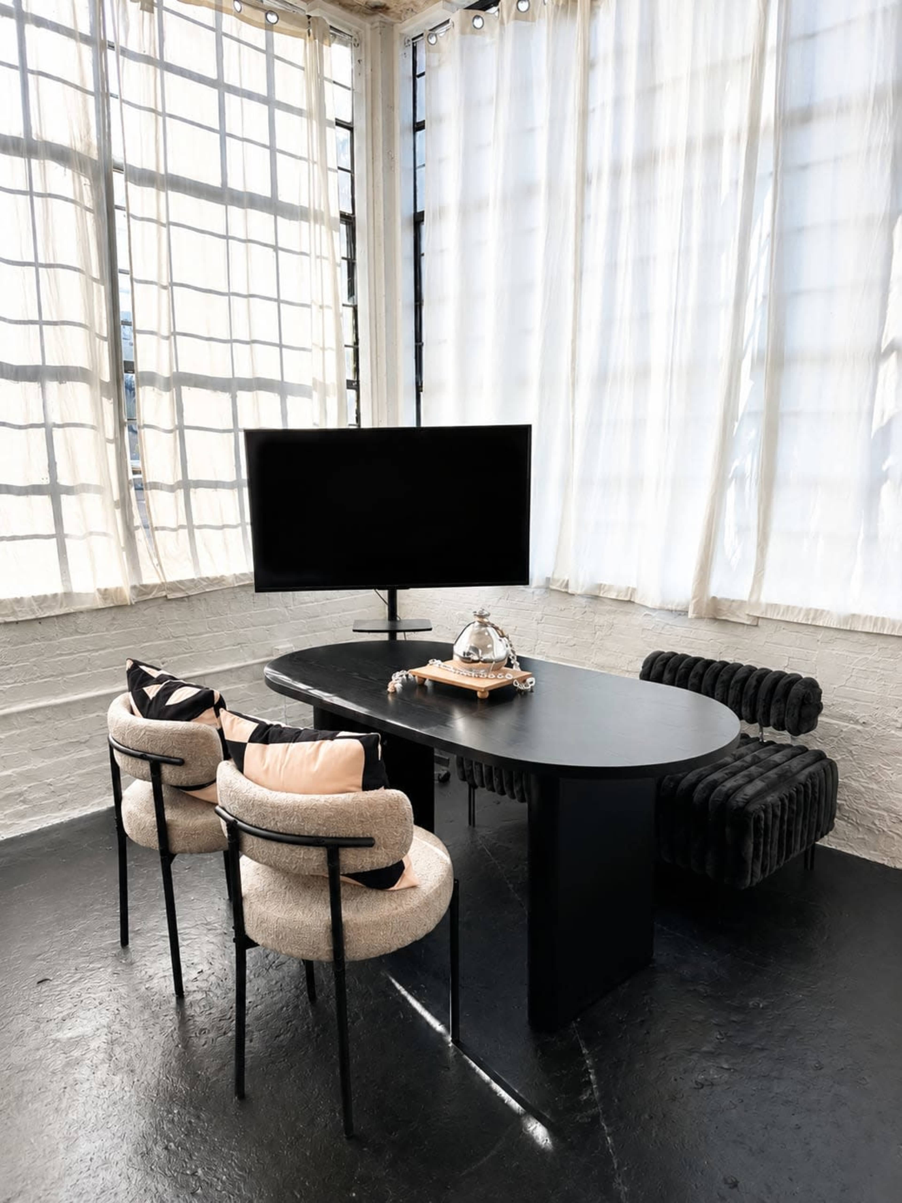 A modern workspace features a black oval table, two stylish chairs, and a television in front of large windows with sheer curtains.