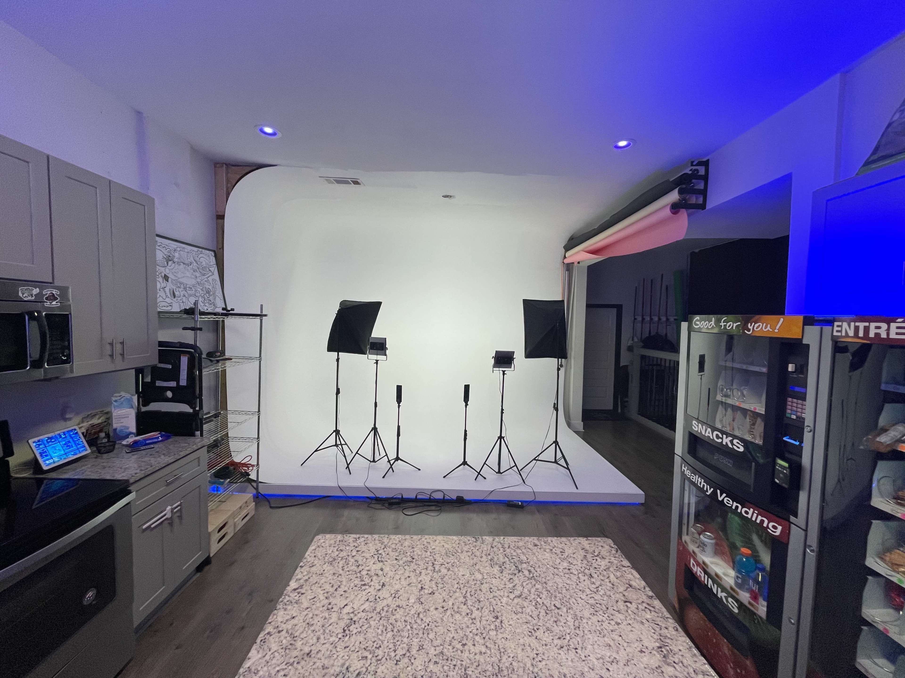 A modern studio space with a white backdrop, two lighting setups, and a kitchen area featuring a vending machine and snack options.