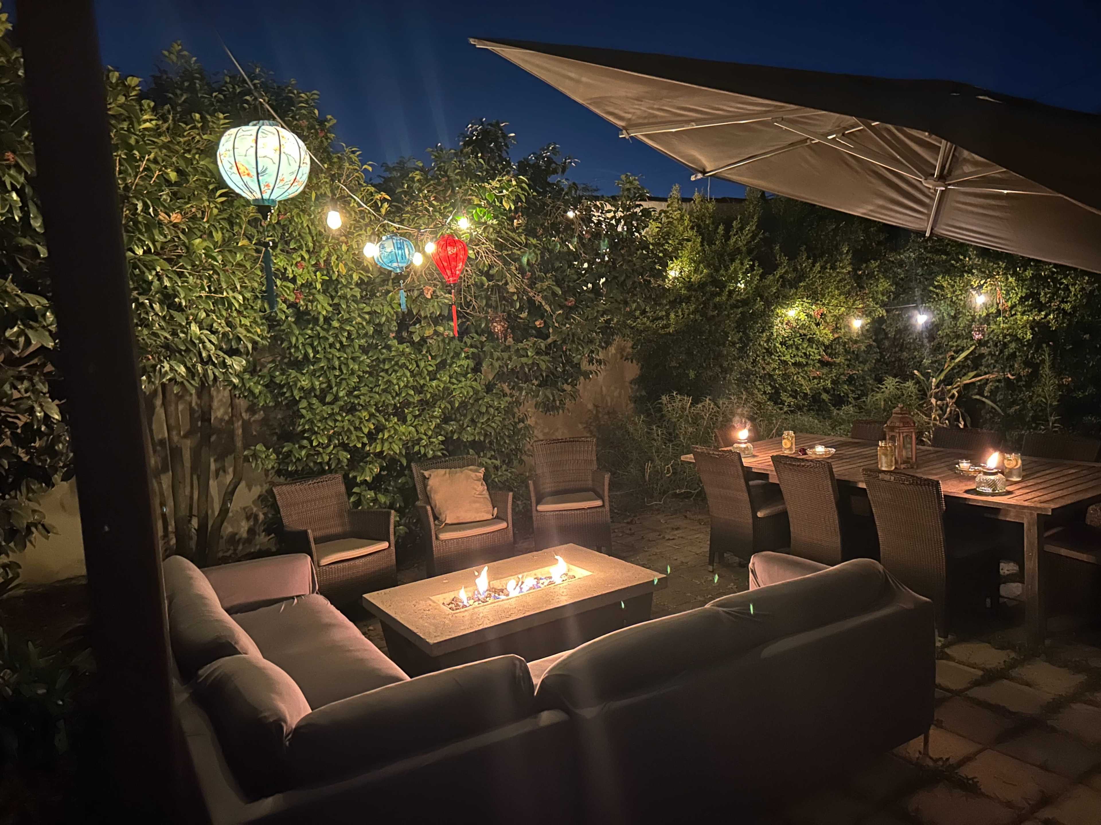A cozy outdoor seating area with a fire pit, surrounded by lush greenery and illuminated by hanging lanterns and string lights at night.