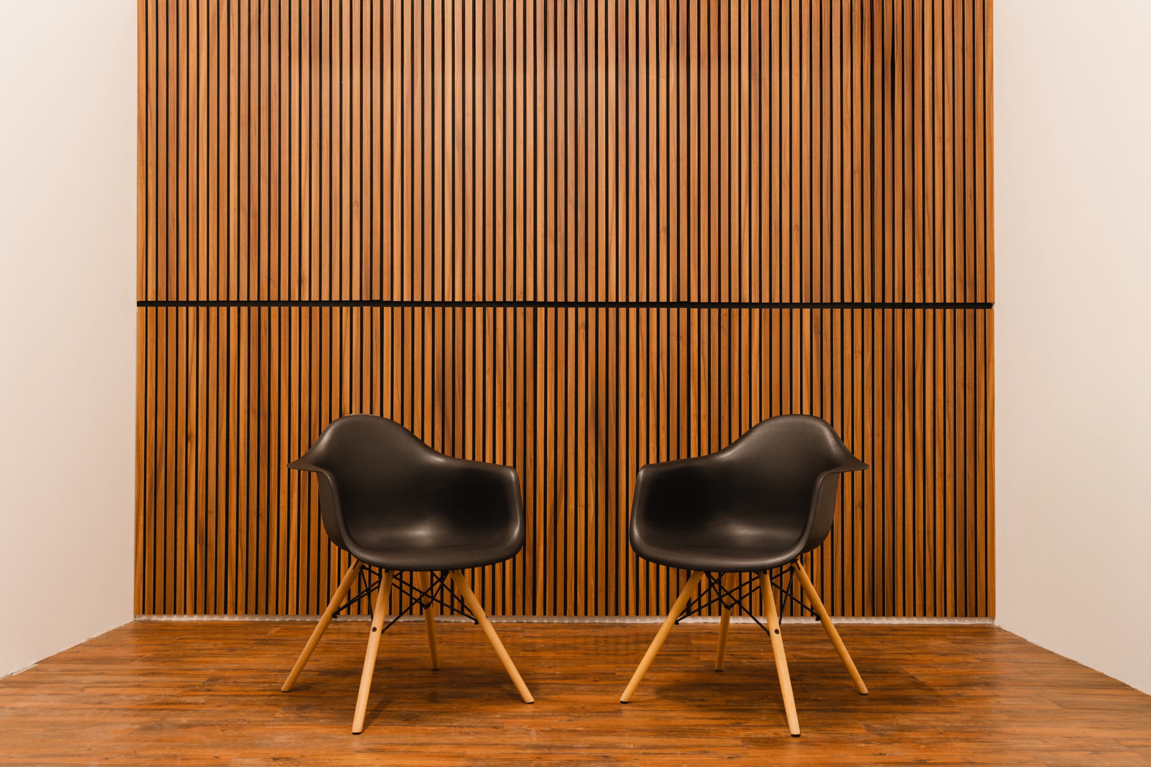 Two black chairs with wooden legs are positioned facing each other in front of a wooden slatted wall.