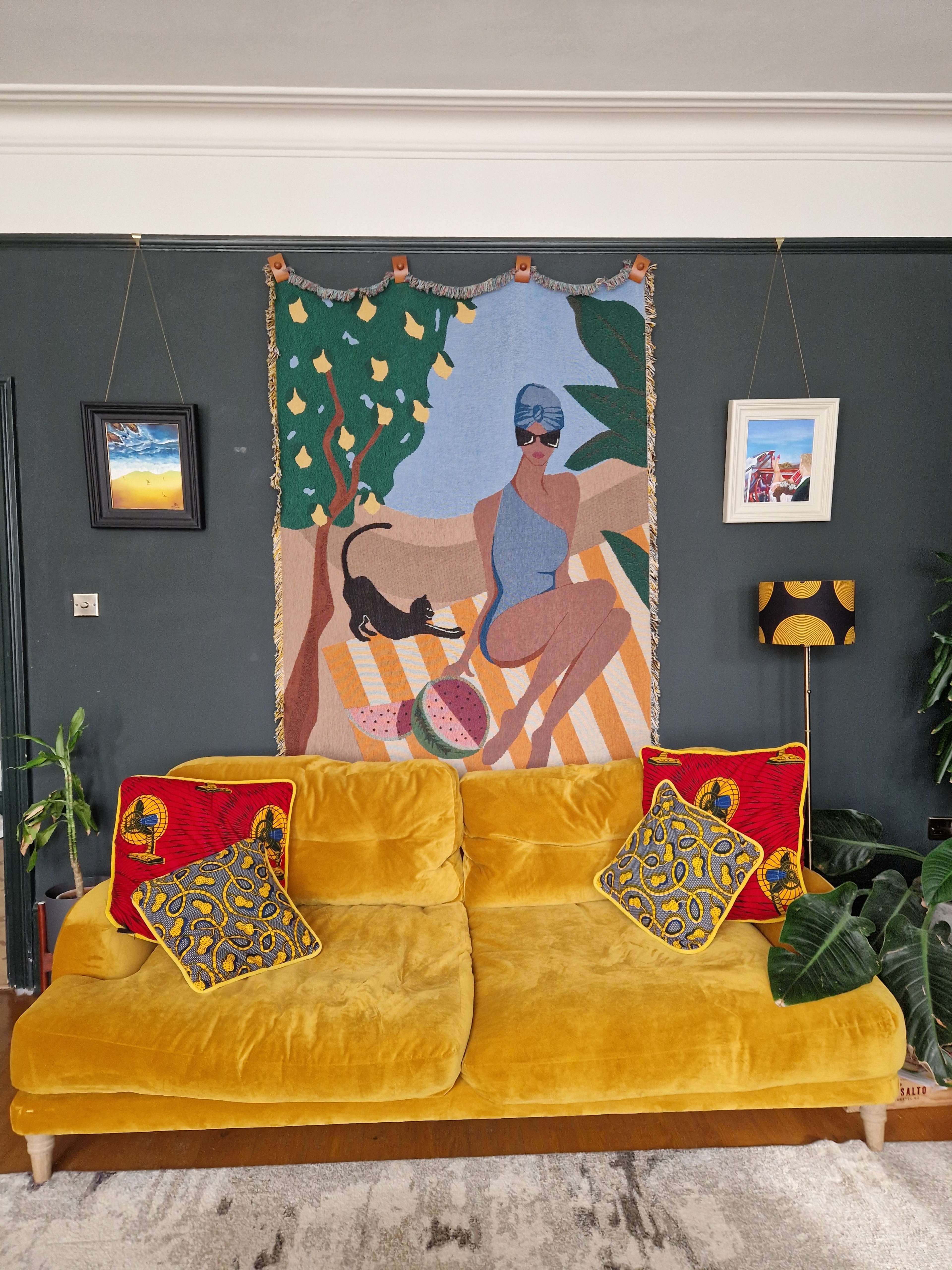 A yellow velvet sofa is adorned with patterned cushions and placed against a dark green wall featuring a colorful tapestry and framed artwork.