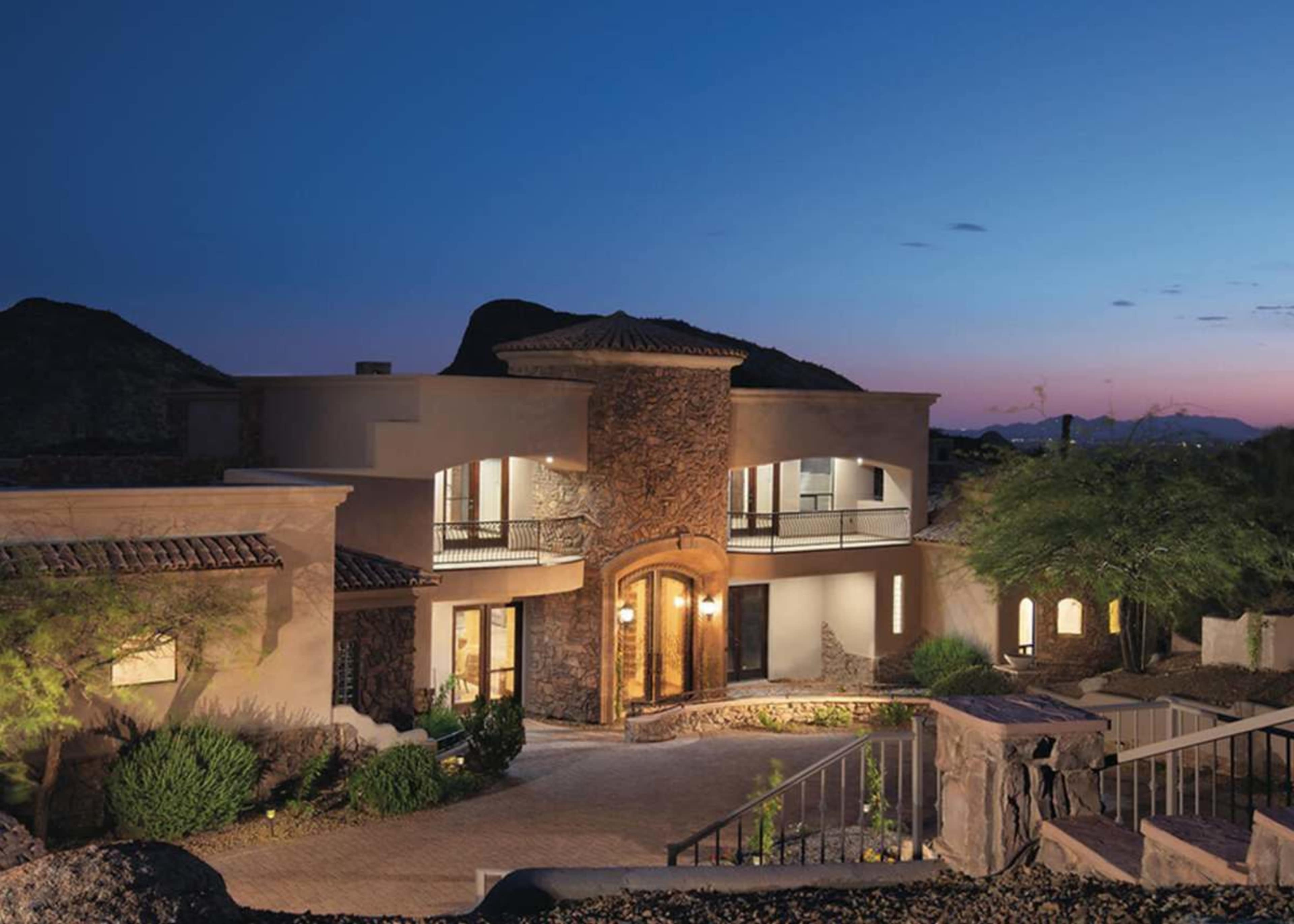The image shows a modern desert home with stone accents and a landscaped driveway, set against a twilight sky.