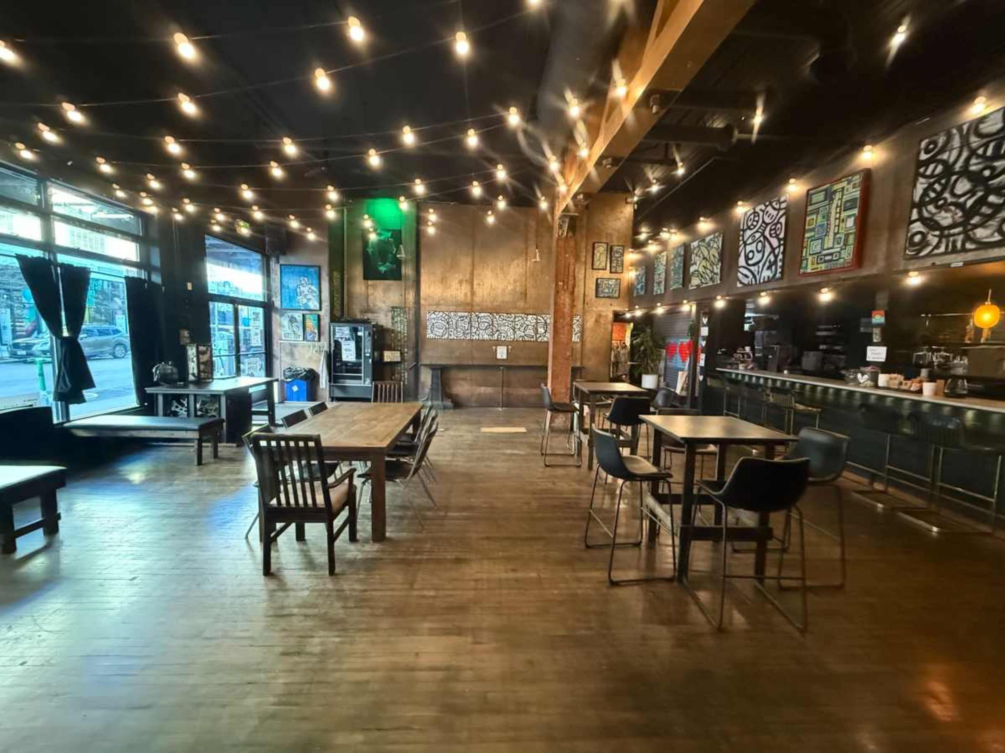 The image shows a spacious café interior with wooden tables, chairs, and a bar area, illuminated by string lights.
