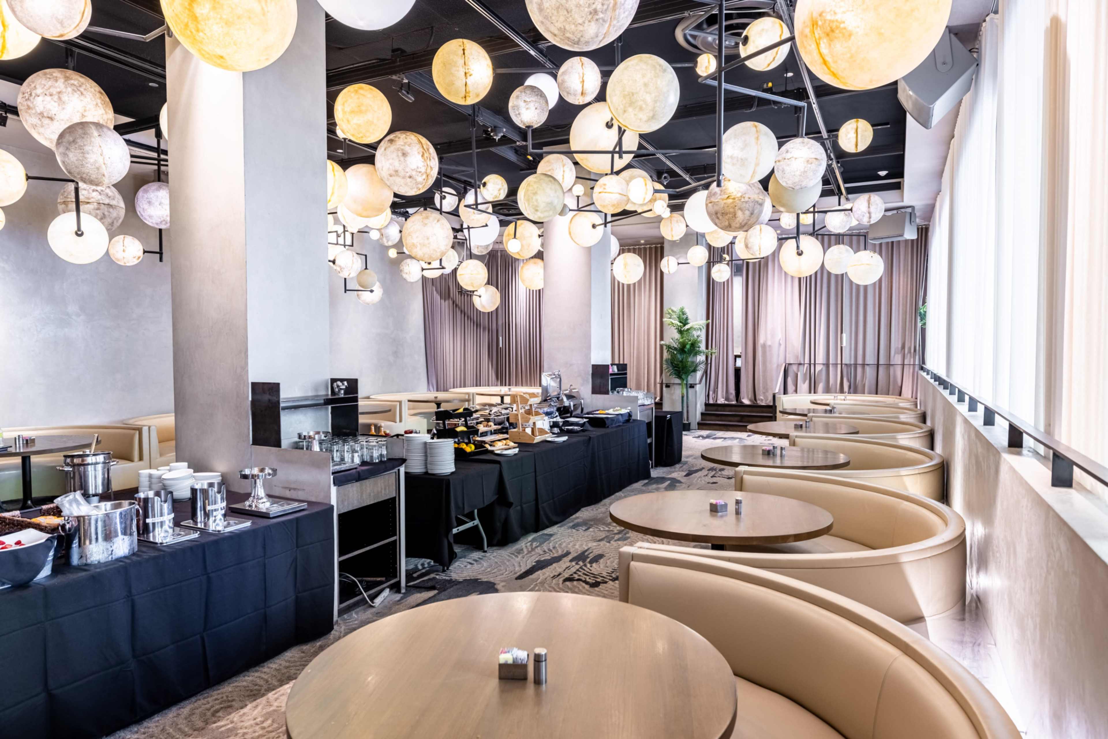 The image shows a modern dining area with round tables, plush seating, and an assortment of spherical light fixtures hanging from the ceiling, alongside a buffet table displaying various dishes.