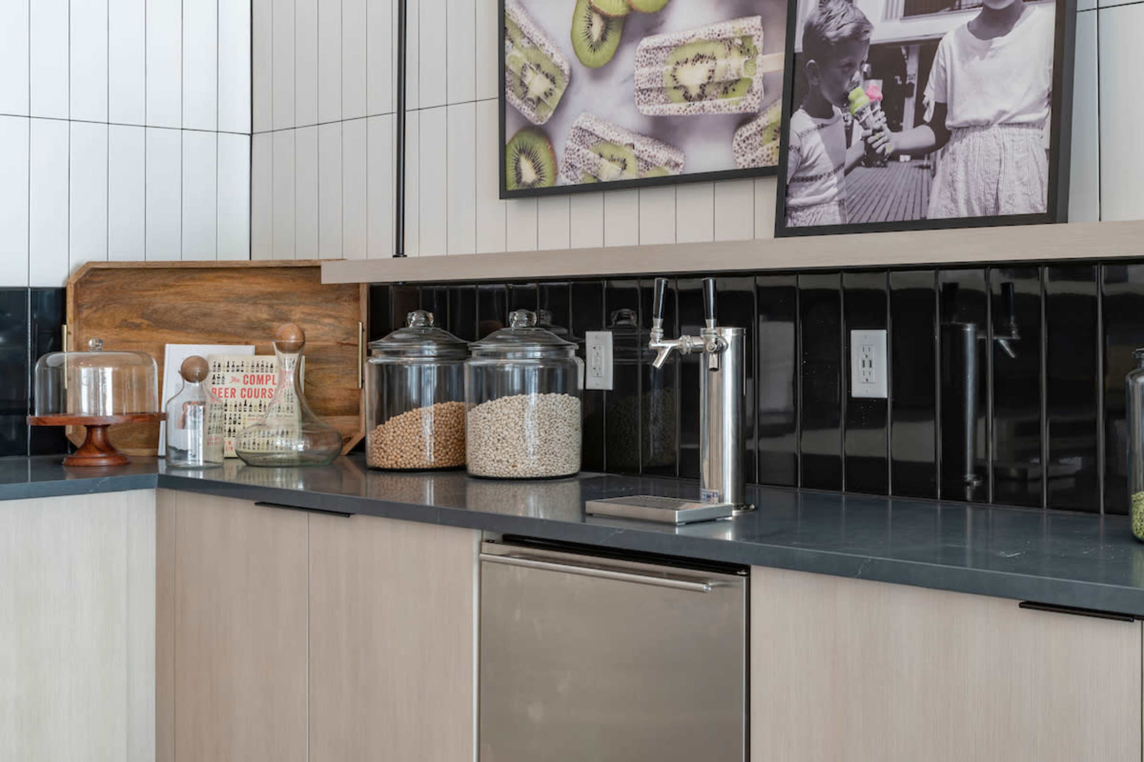 The image displays a modern kitchen with a countertop featuring glass jars of grains, a beverage dispenser, and a stainless steel dishwasher beneath a black and white photo collage.