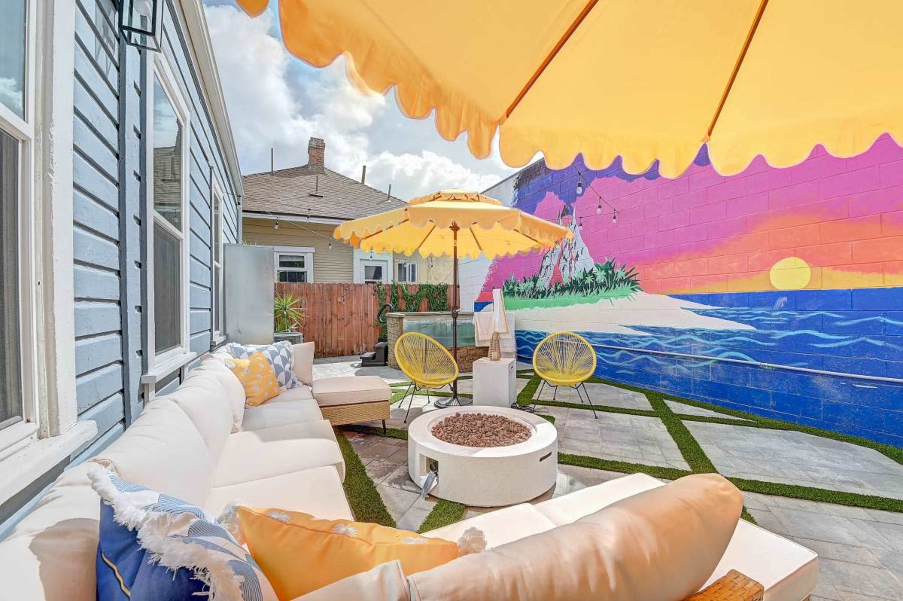 A colorful outdoor seating area features a sectional couch and yellow umbrellas next to a vibrant mural of a beach scene.