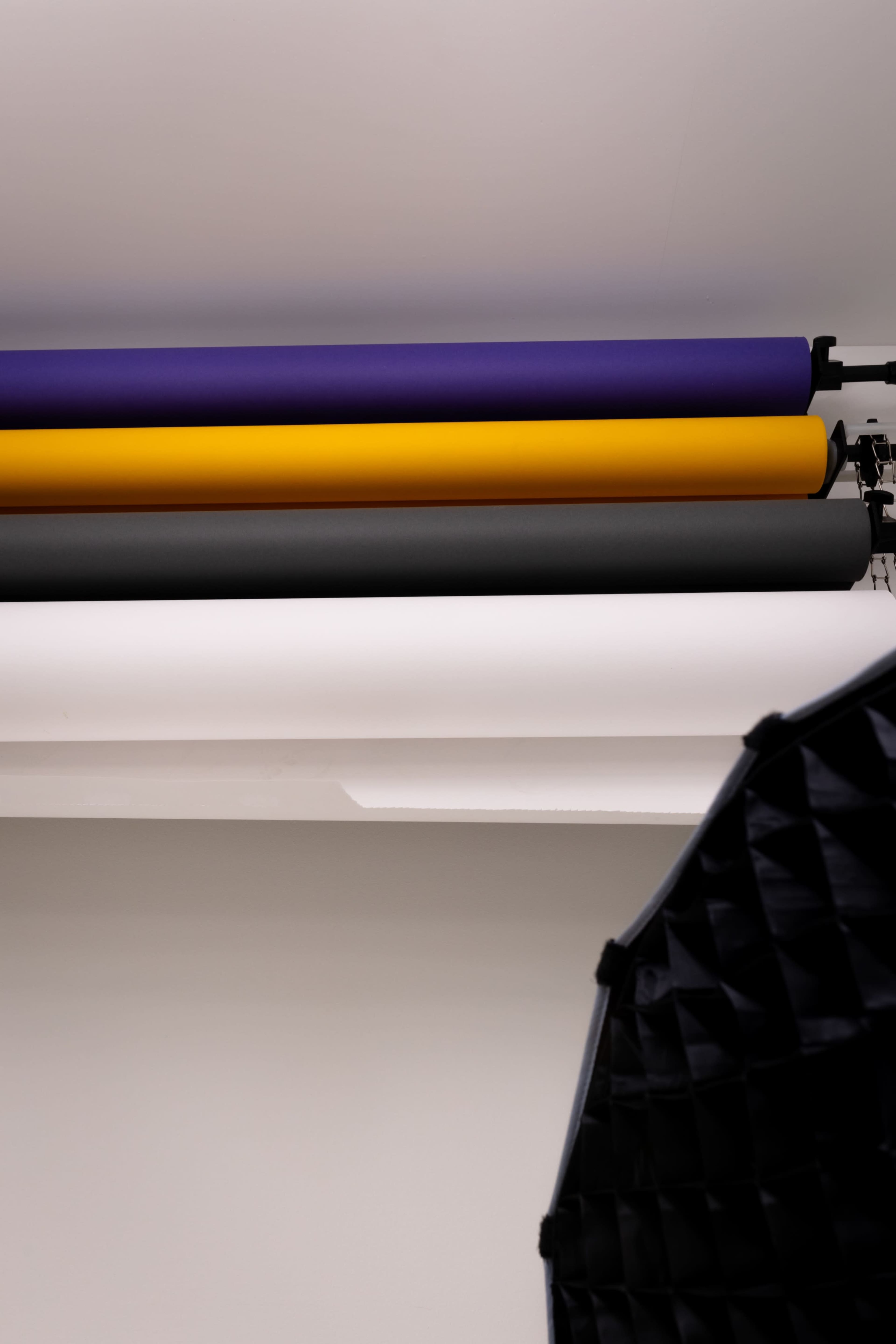 The image shows a backdrop setup with rolls of paper in purple, yellow, white, and gray, along with a black light modifier in the foreground.