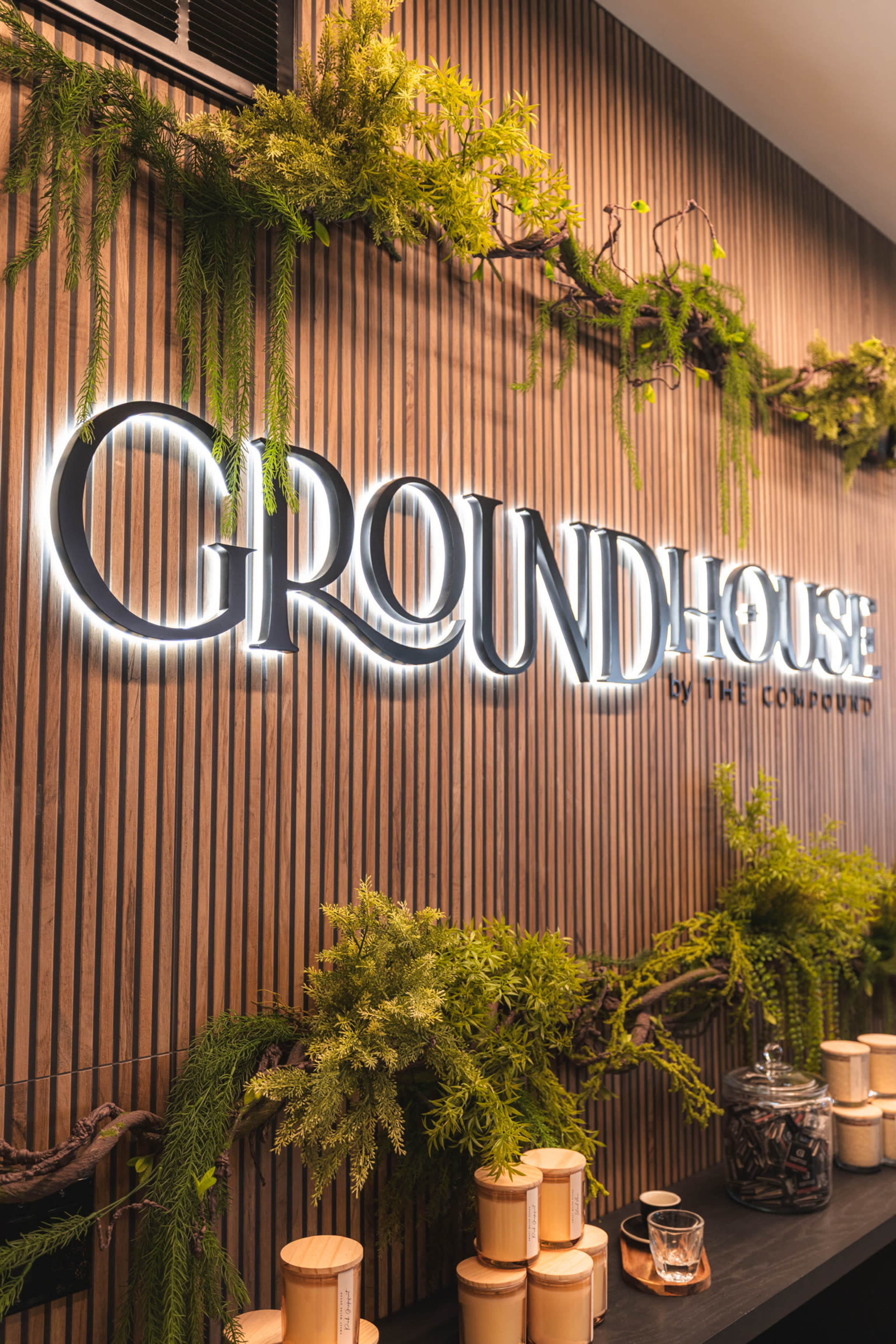 The image shows a modern interior with a wooden wall featuring the illuminated sign "GROUNDHOUSE by THE COMPOUND" surrounded by greenery.