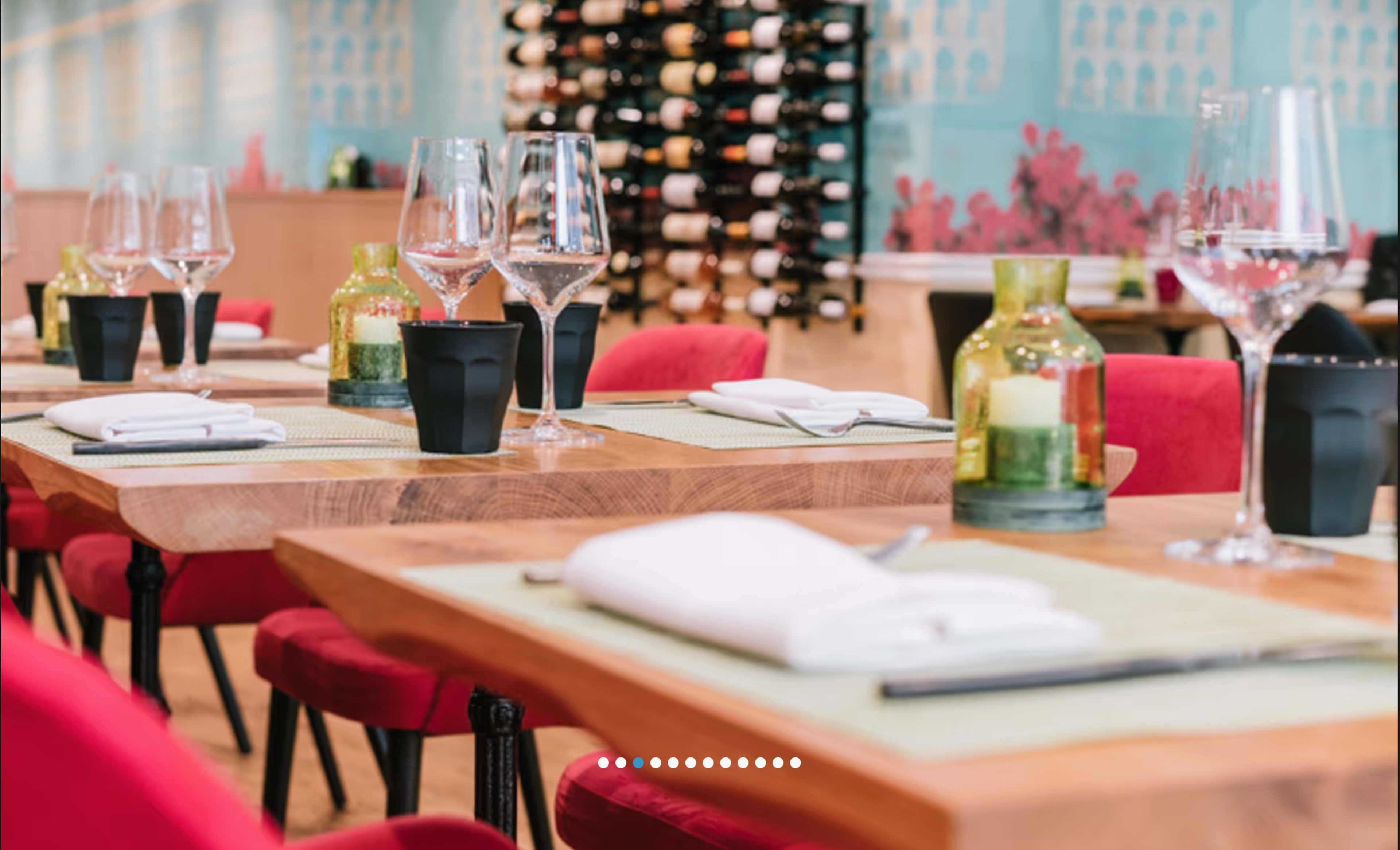 The image shows a stylish restaurant interior with wooden tables set with glasses, napkins, and decorative centerpieces, surrounded by red chairs and a wine display on the wall.