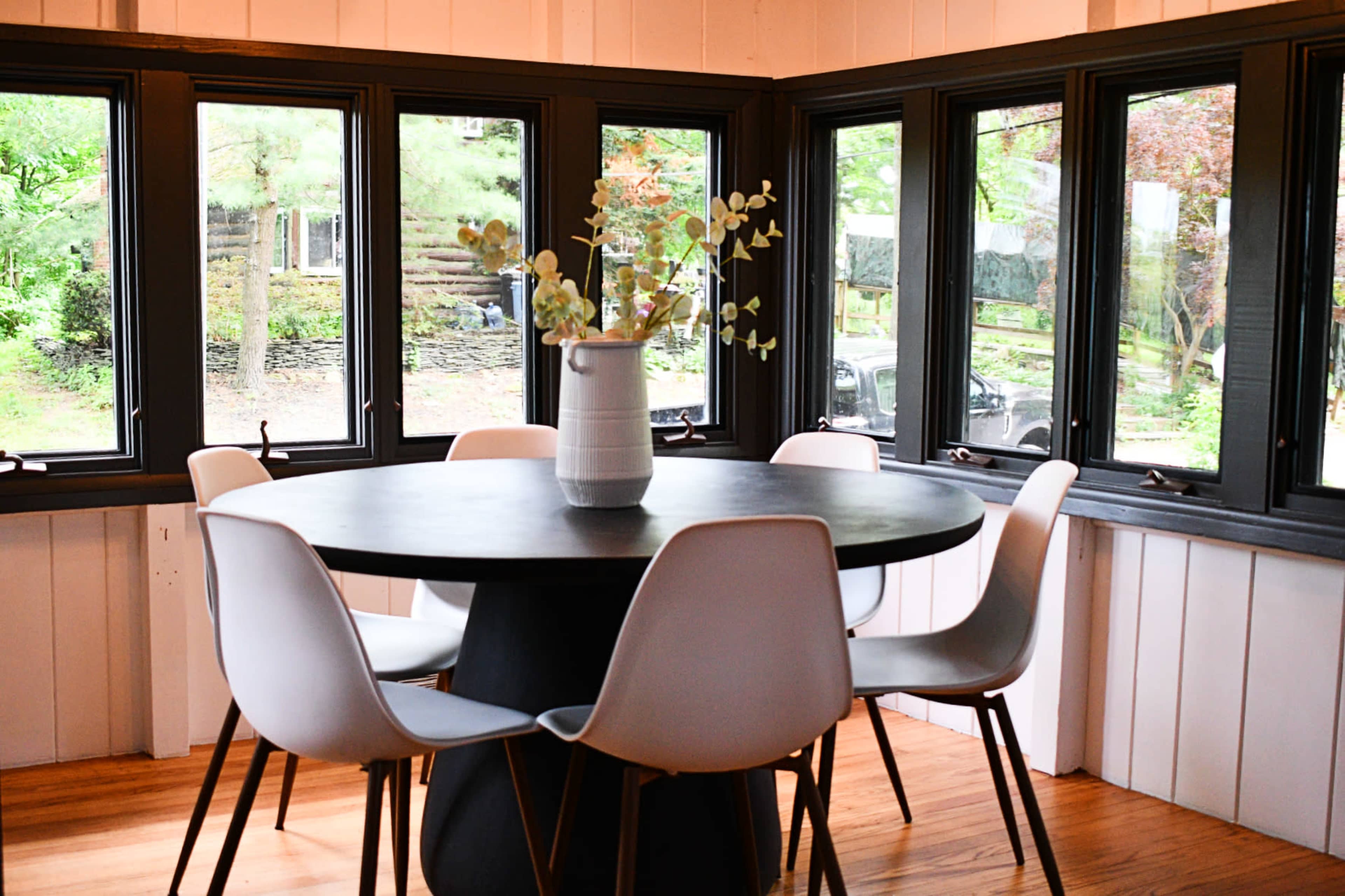 A round black table surrounded by white chairs in a bright room with large windows overlooking a wooded area.