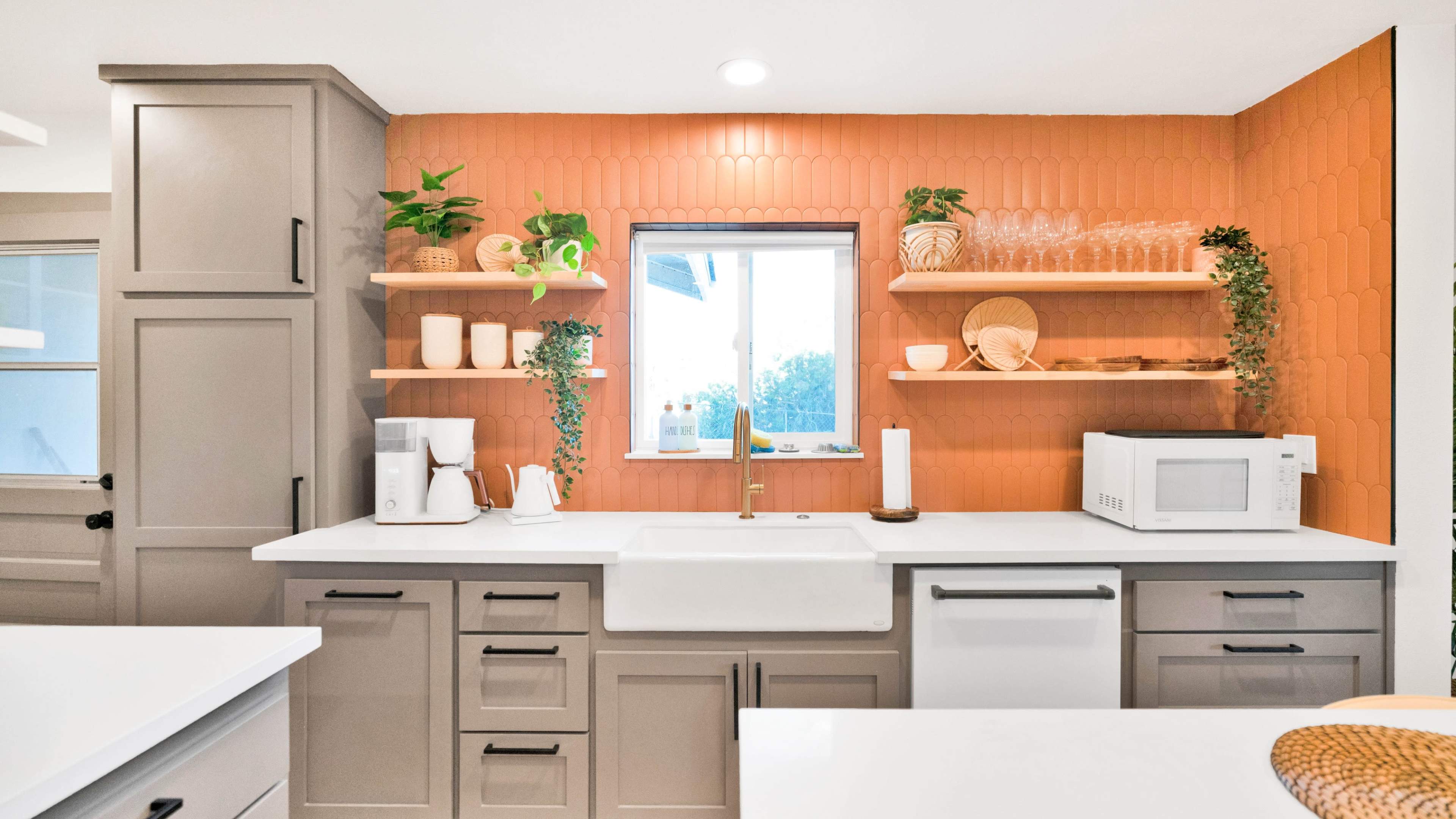 A modern kitchen features a white sink and countertop with gray cabinets, open shelves, and an orange tiled backsplash.