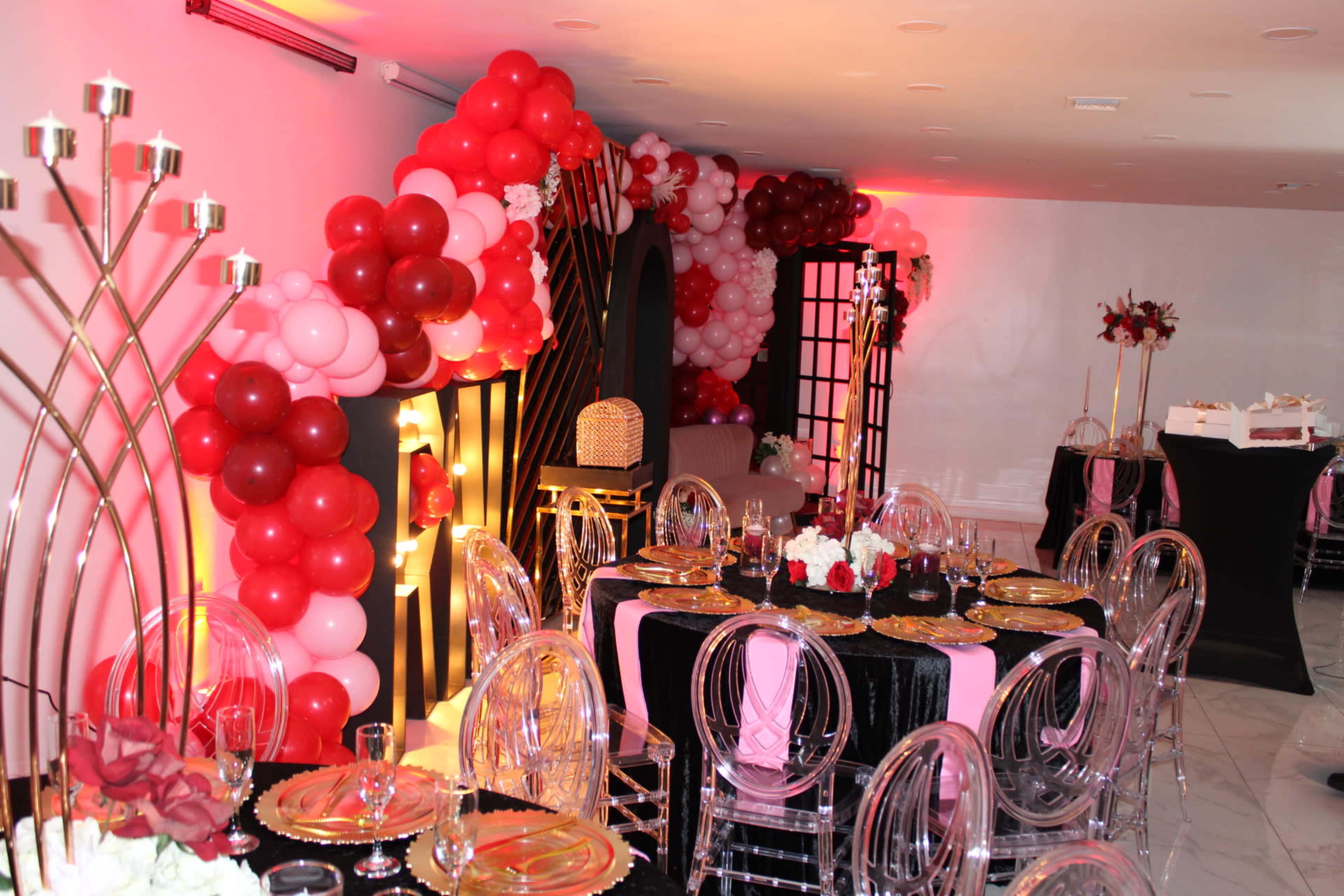 A decorated event space features an arrangement of black and pink tables with transparent chairs, adorned with floral centerpieces and balloon archways in red and pink.