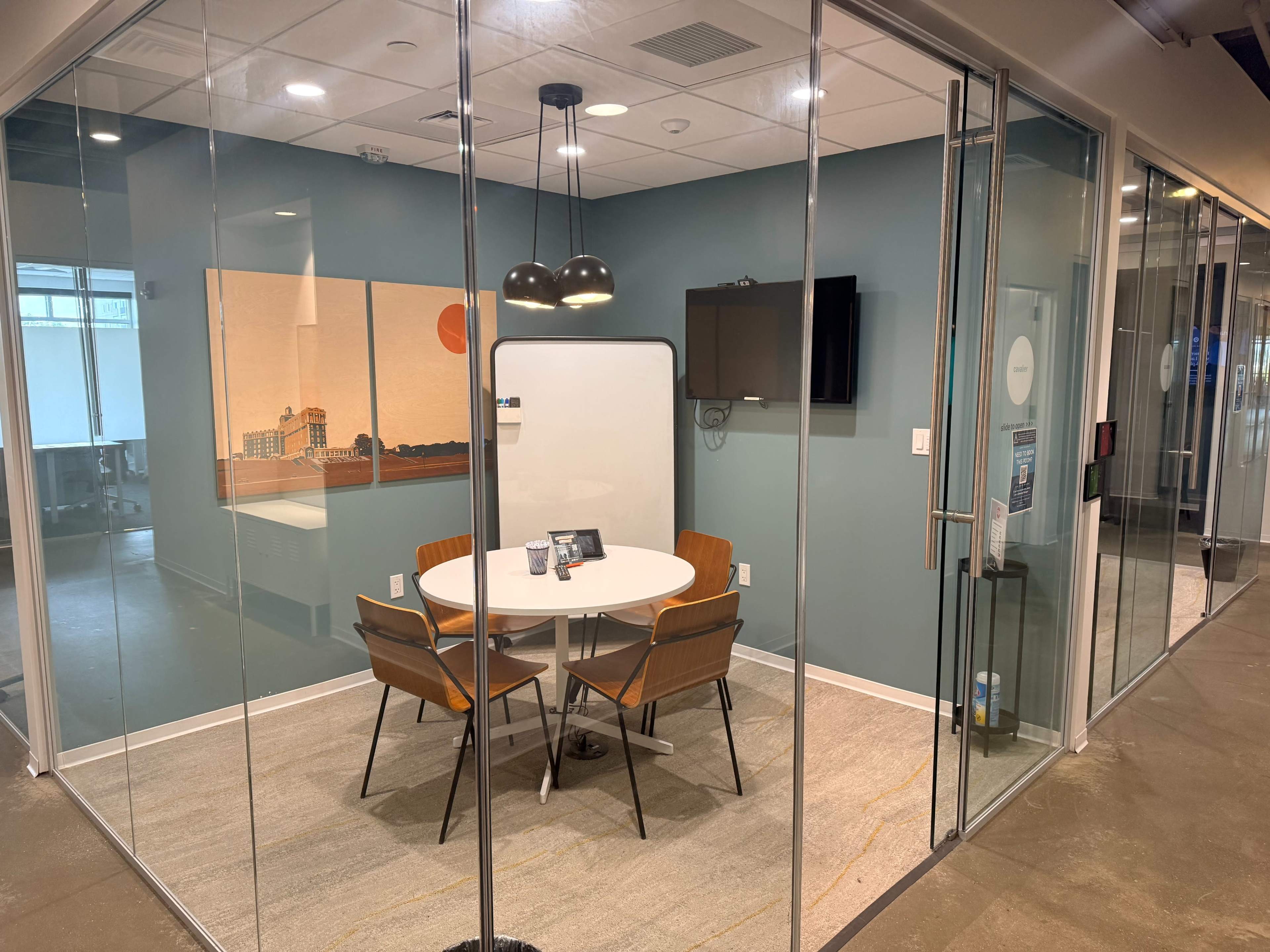 The image shows a glass-walled conference room with a round table and four chairs, featuring a TV and a whiteboard on the wall.